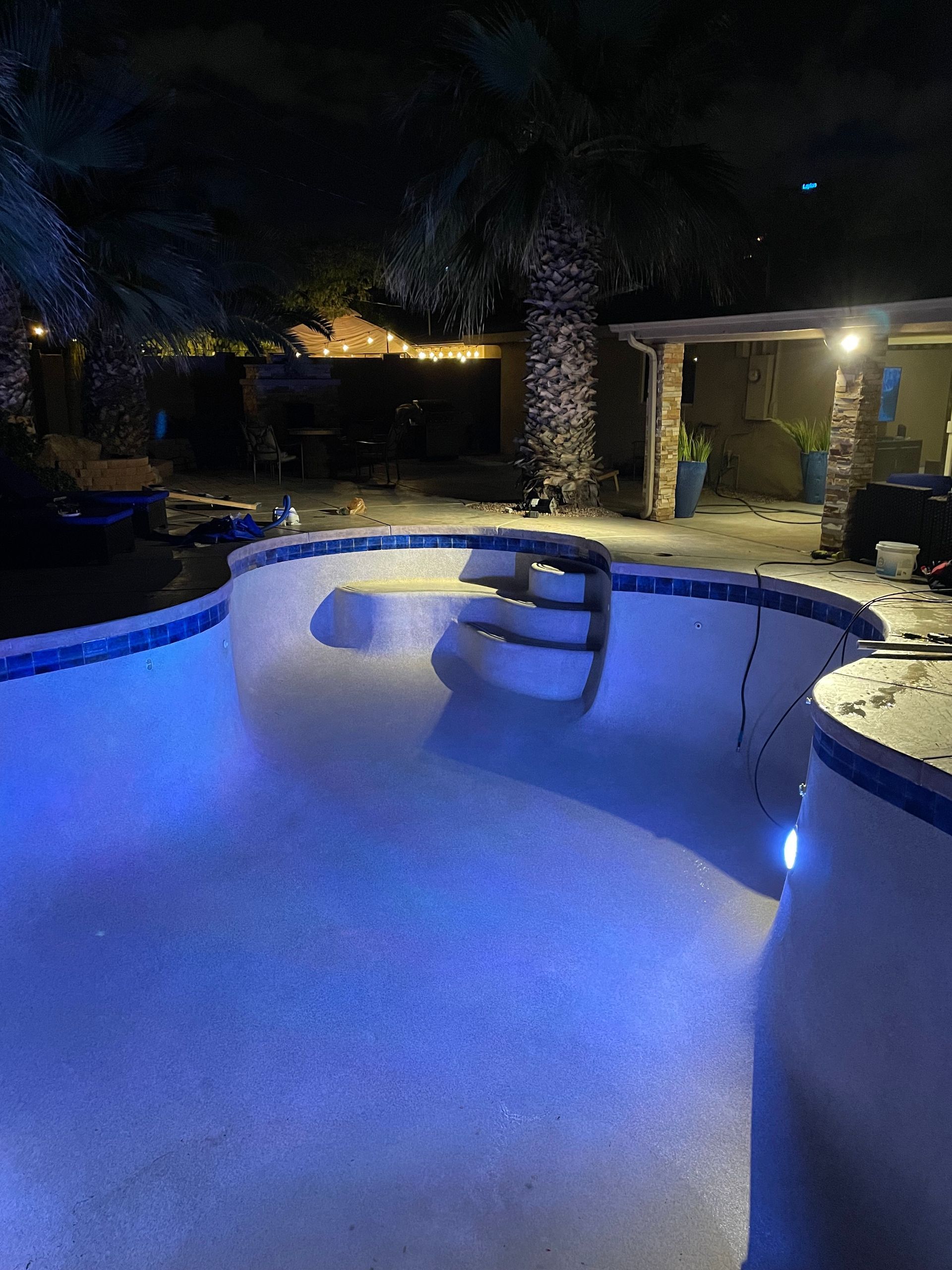 A nighttime view of an empty pool illuminated by blue lights, with a palm tree and a covered patio in the background.