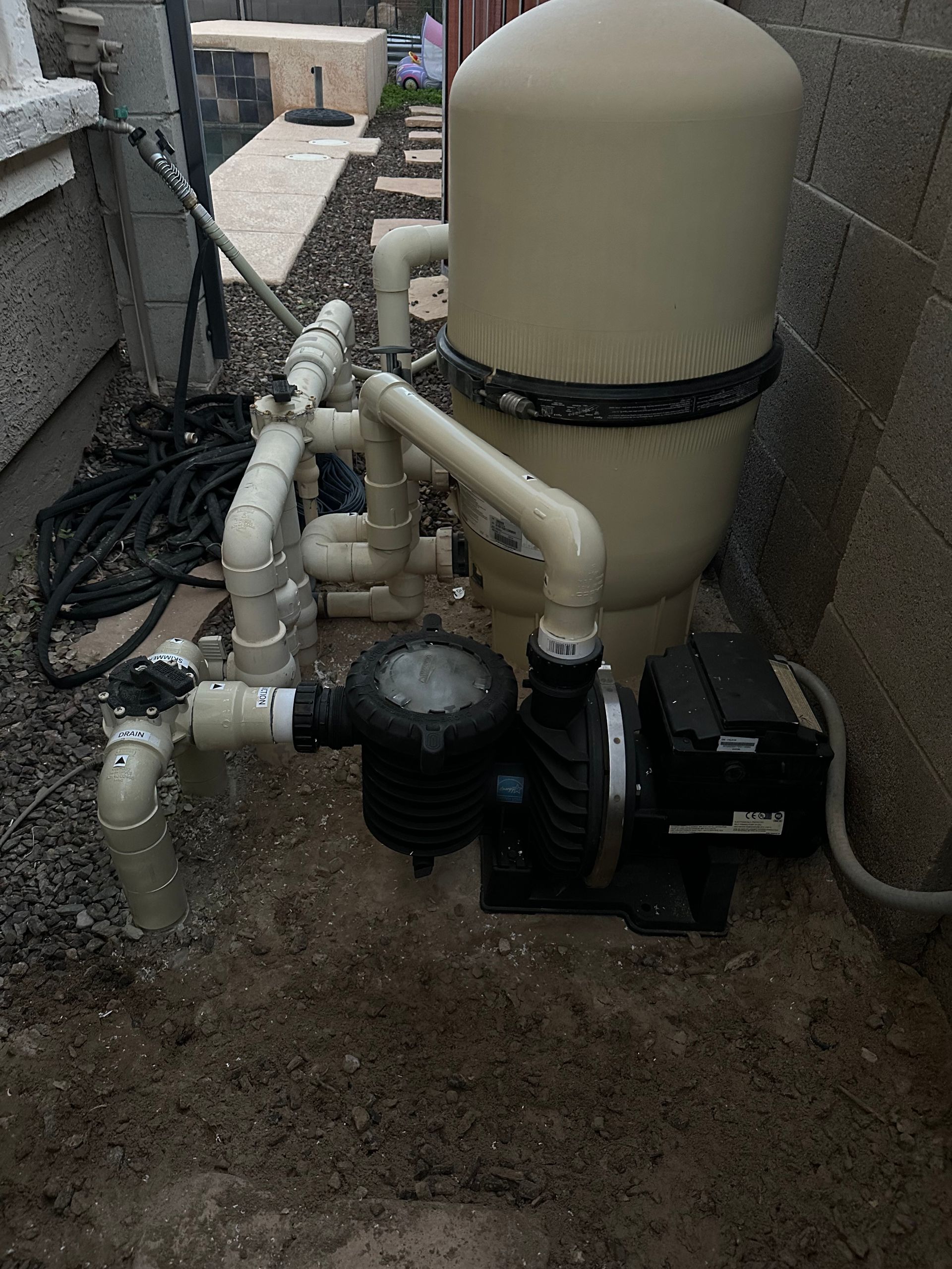 Pool pump and filter system installed outdoors, beige tank, black pump, connected by white PVC pipes.