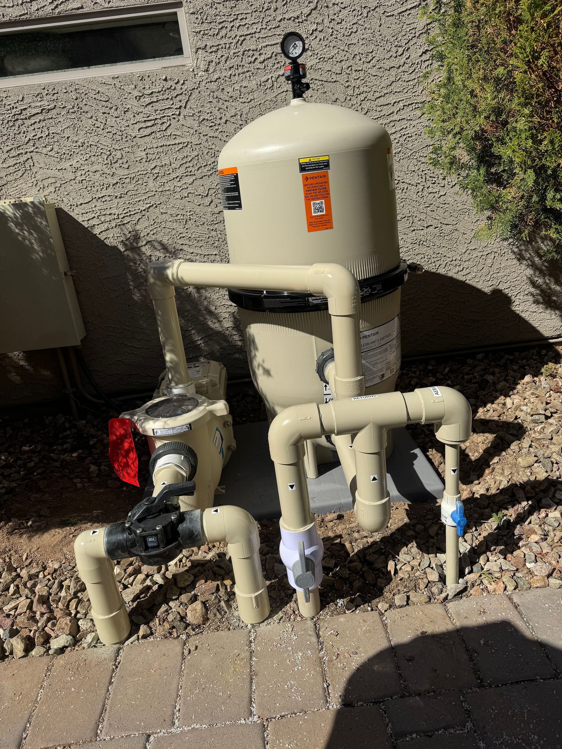 A tan pool filter system with plumbing on a gravel and brick patio next to a stucco wall.