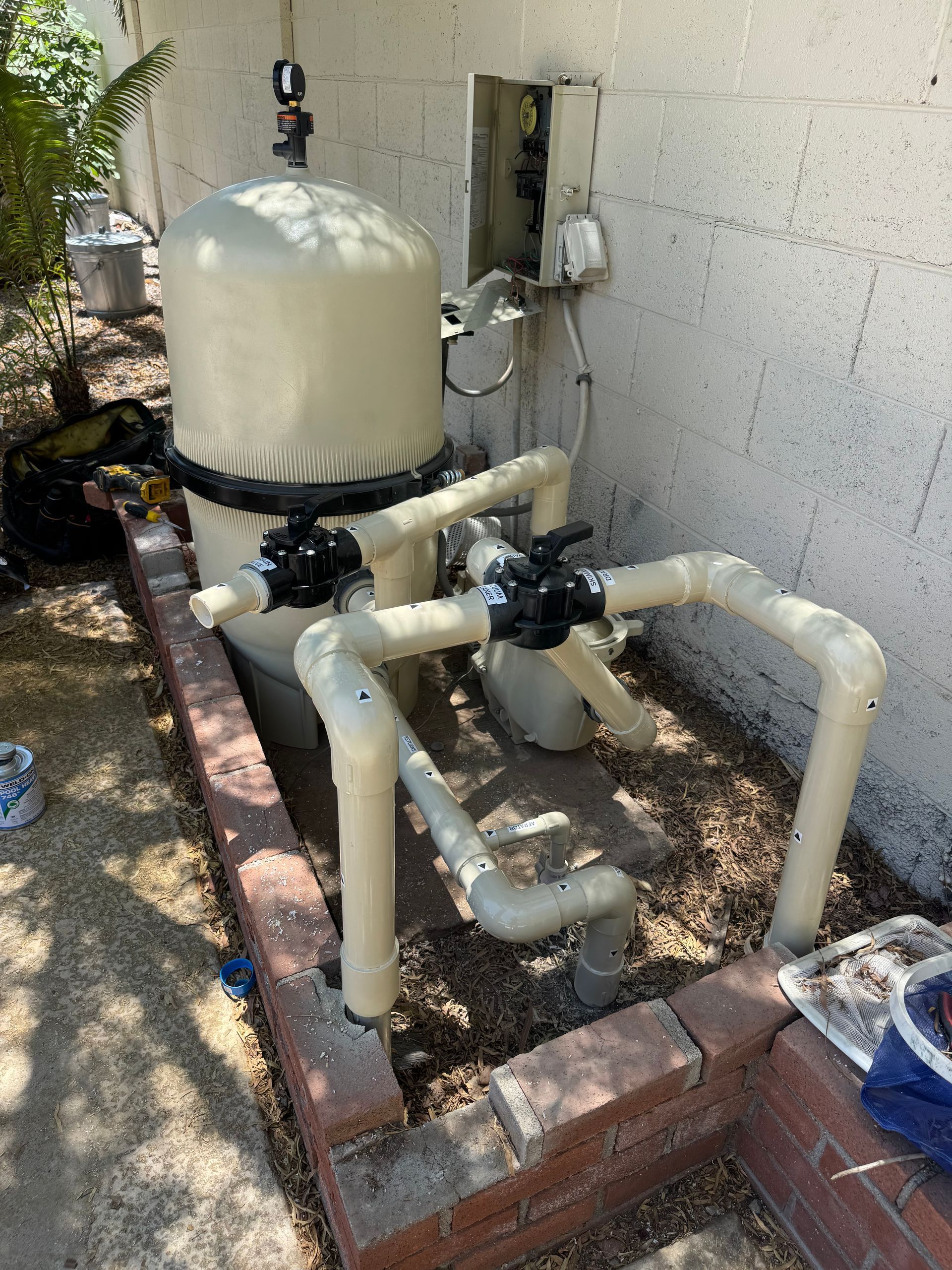 Pool filtration system with beige tank, pipes, and valves near a white wall.