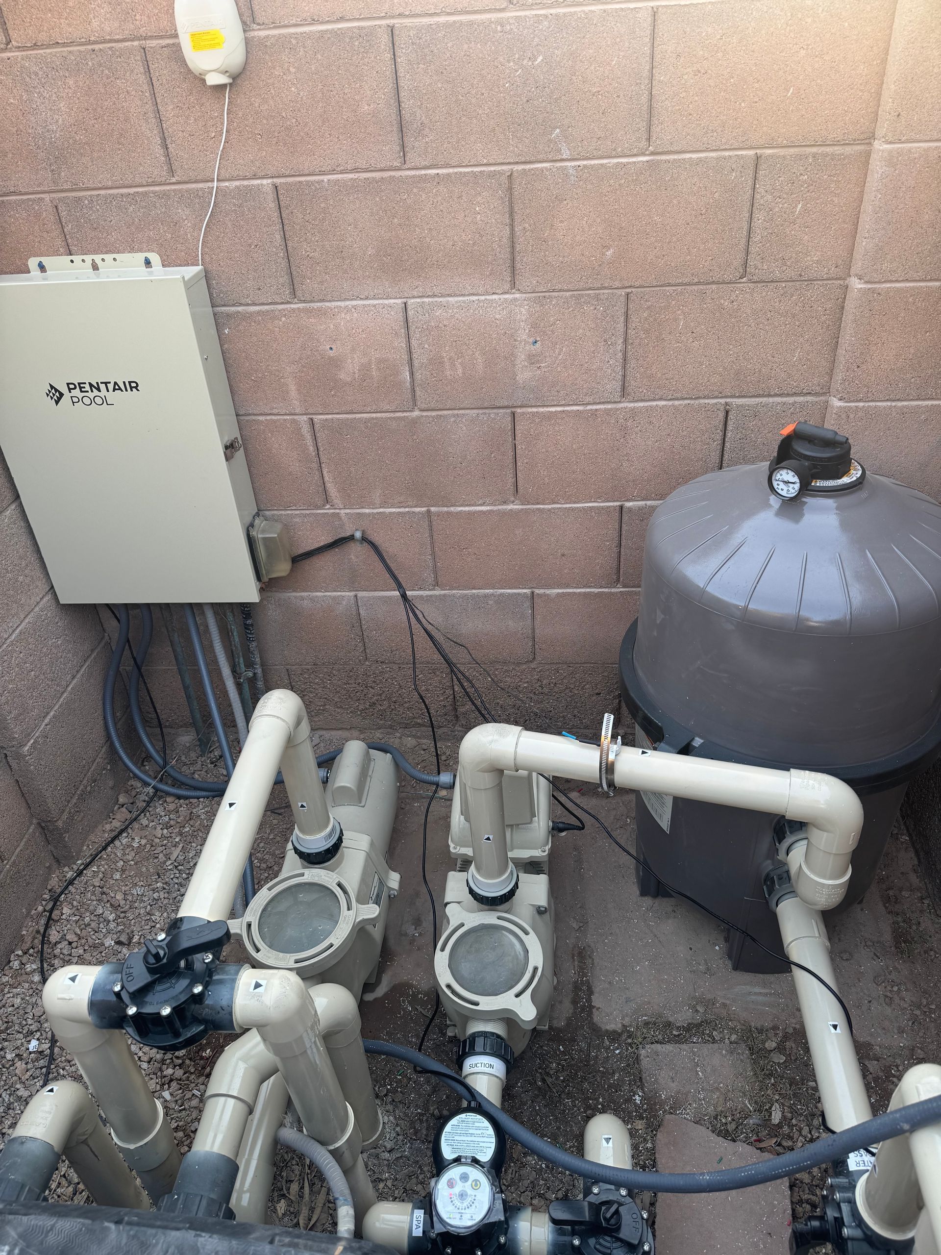 Pool equipment, including pumps, filter, and control box, against a textured brick wall in an outdoor setting.