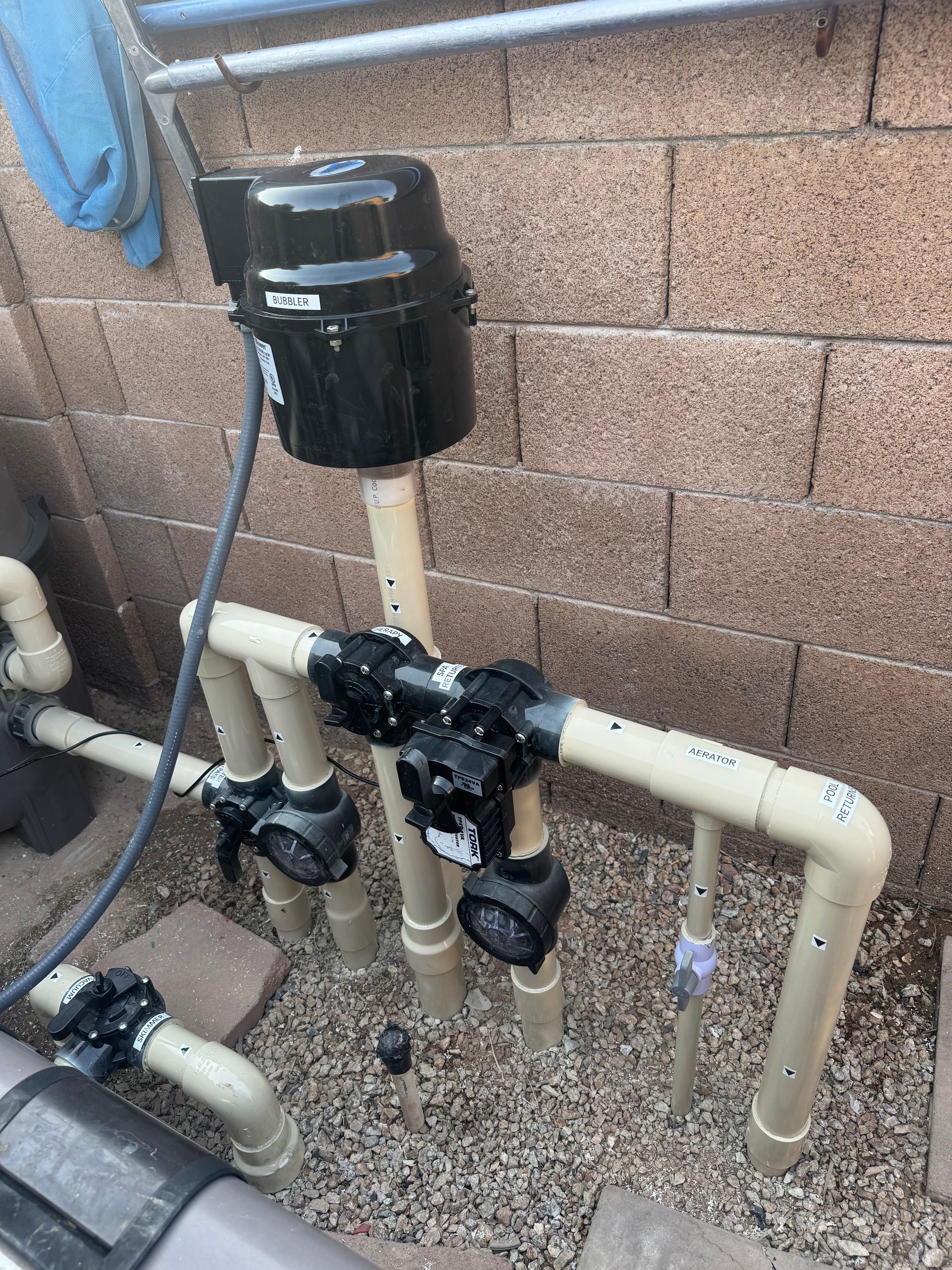 Pool equipment: beige pipes, black valves/motor, against a brick wall, outdoors.