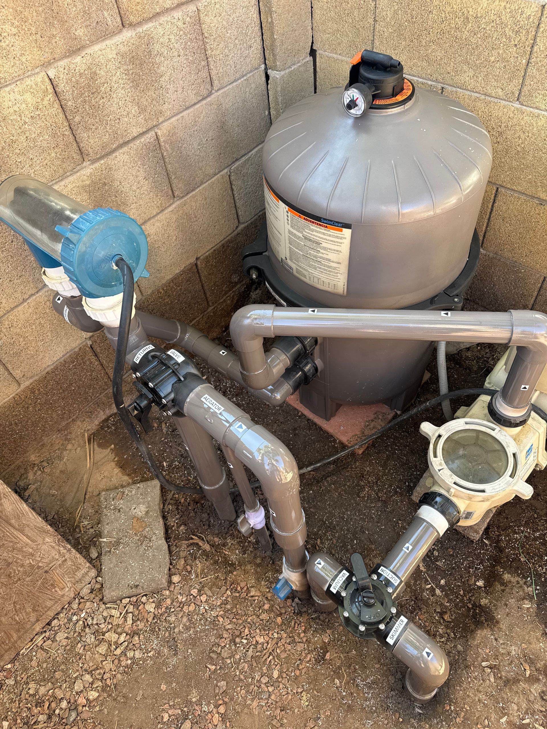 Pool filter system with gray tank, pipes, and pump against a tan brick wall.