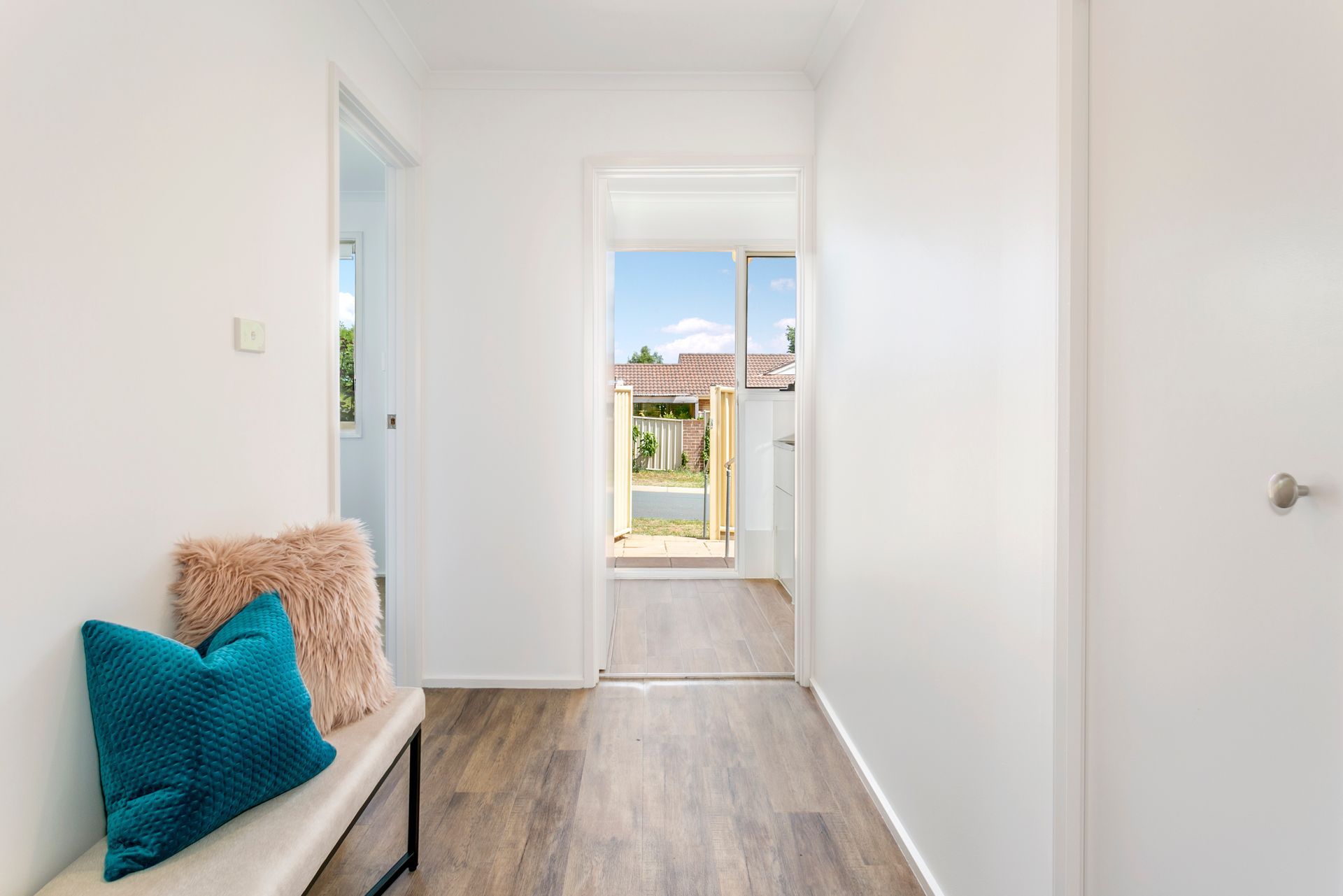 Hallway with wooden floor, white walls, bench with pillows. View to doorway and outside.