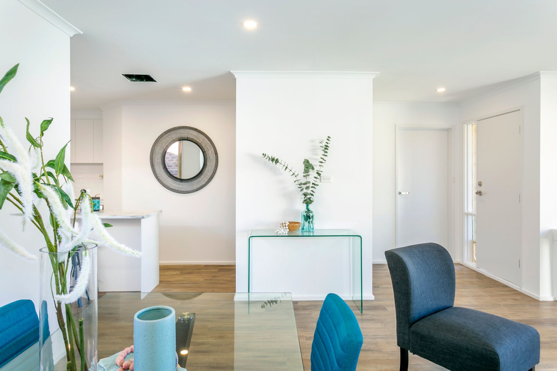 Bright dining room with a glass table, blue chairs, and a glass console table with greenery and a mirror on the wall.