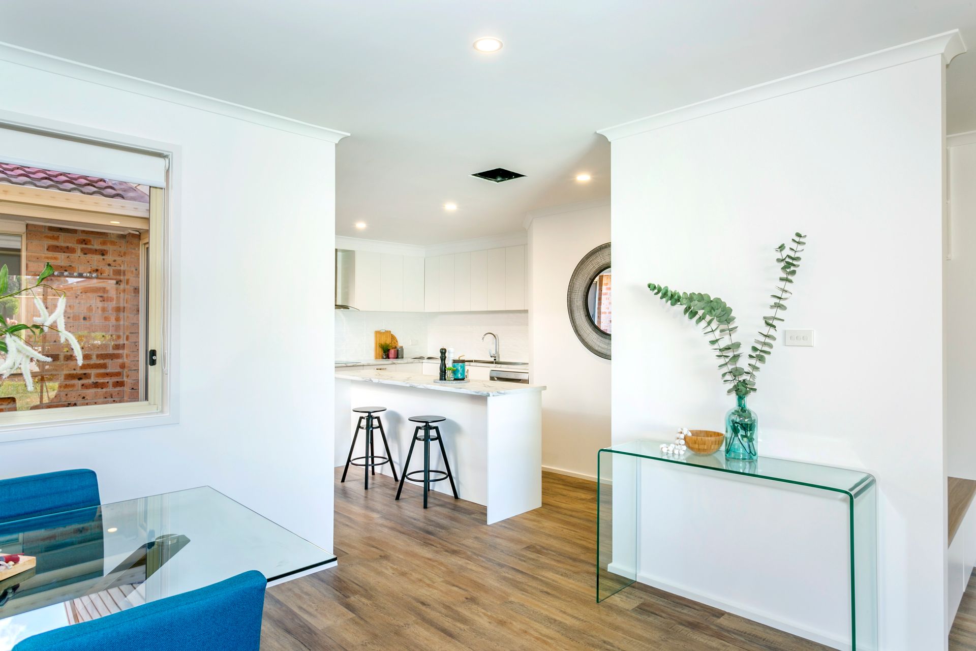 Interior view of a modern apartment with a dining area, kitchen, and glass table. White walls, wood floor, and blue chairs.