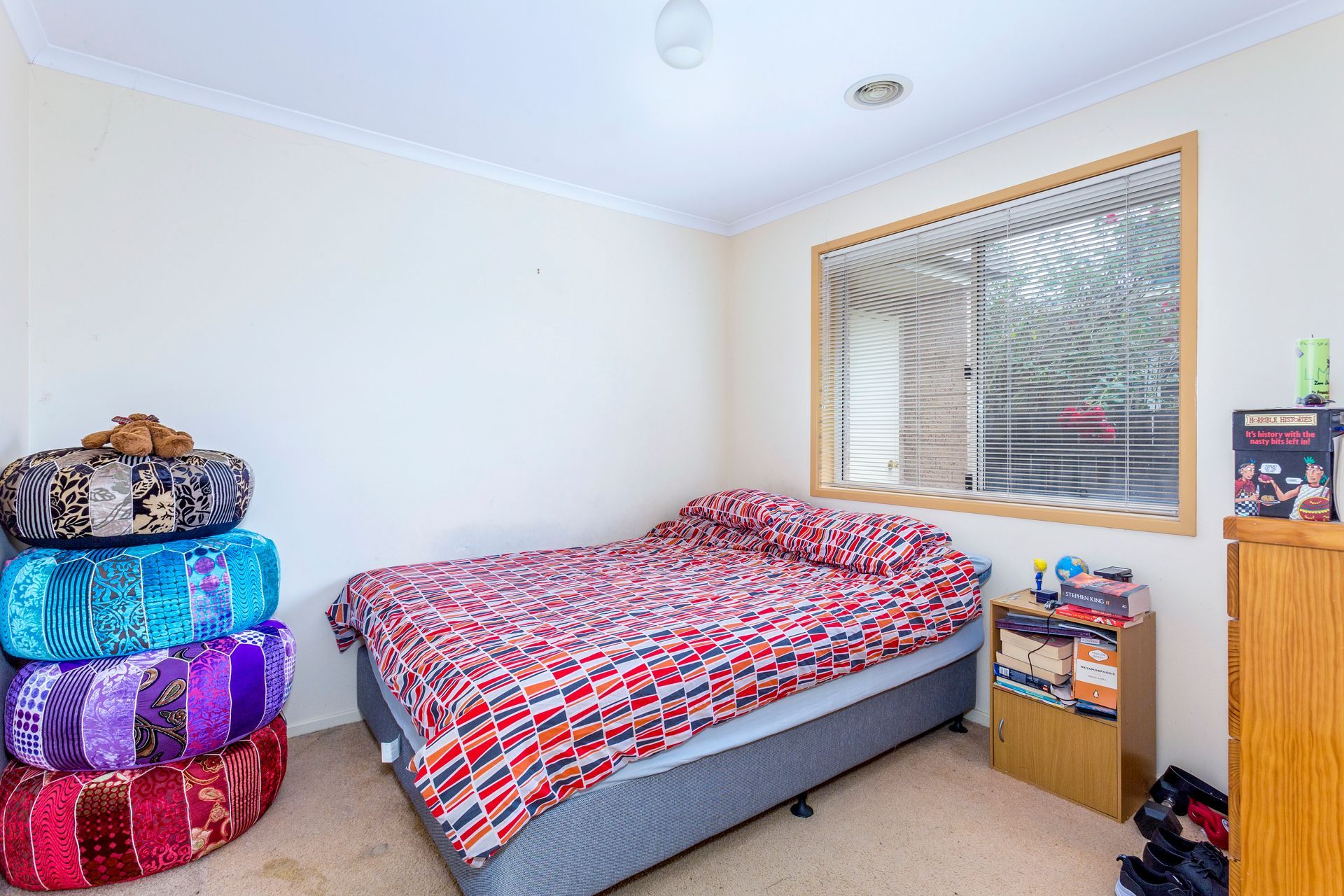 Bedroom with bed, stacked cushions, window, and small cabinet.