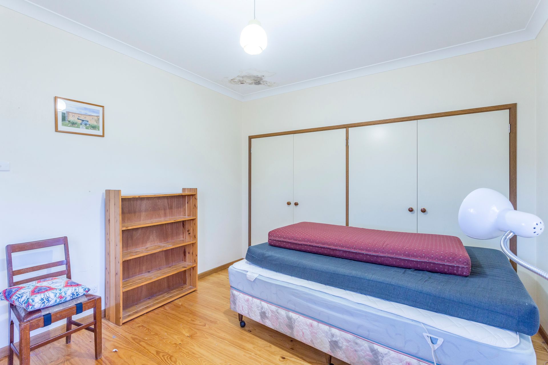 Bedroom with a bed, closet, bookshelf, chair, and floor lamp. Beige walls and wooden floor.