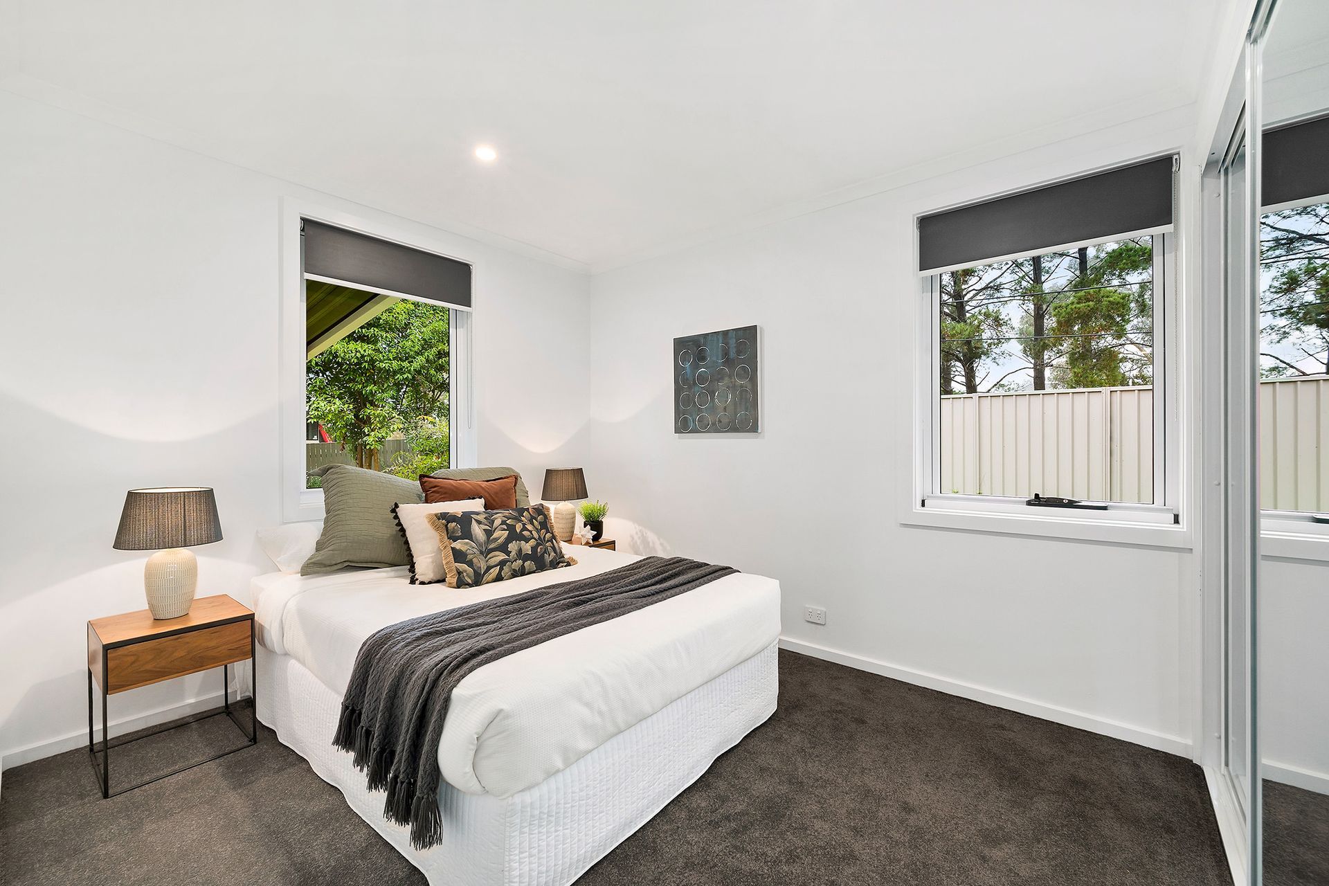 Bedroom with white walls, gray carpet, a bed with pillows and blanket, two windows, and a side table with a lamp.