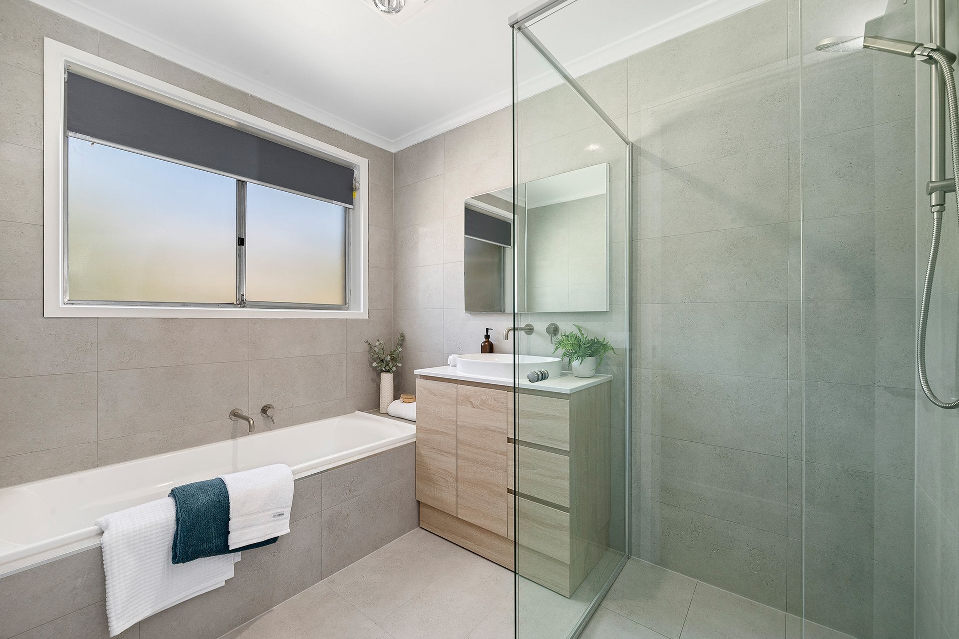Bathroom with a bathtub, shower, vanity, and window with a dark gray blind; gray tiled walls.