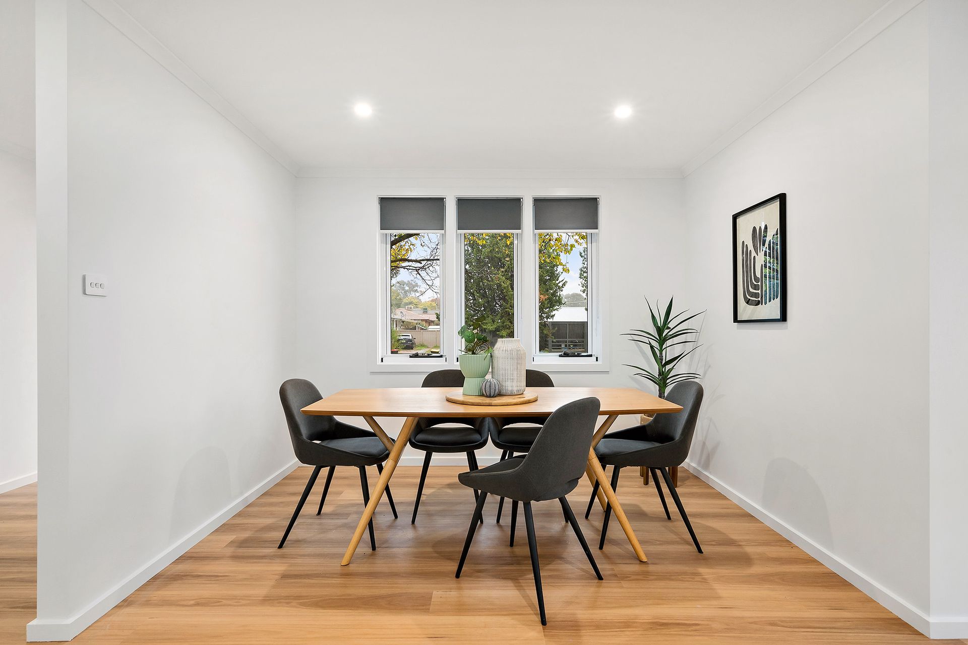 Dining room with wooden table, gray chairs, window, art, and plant. Light wood flooring and white walls.