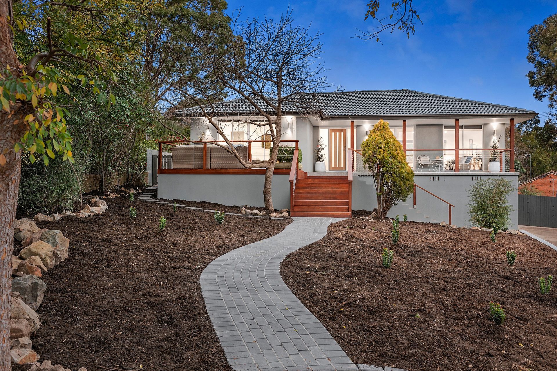 House with gray roof, wooden deck, and winding brick path in a landscaped yard.