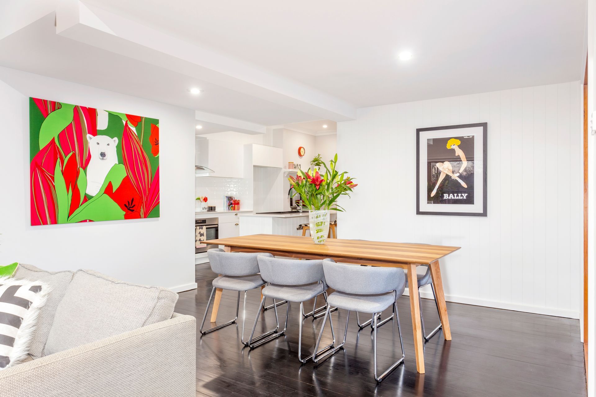 Dining room with table, chairs, art, and kitchen in the background. White walls, dark floors, and a colorful painting.