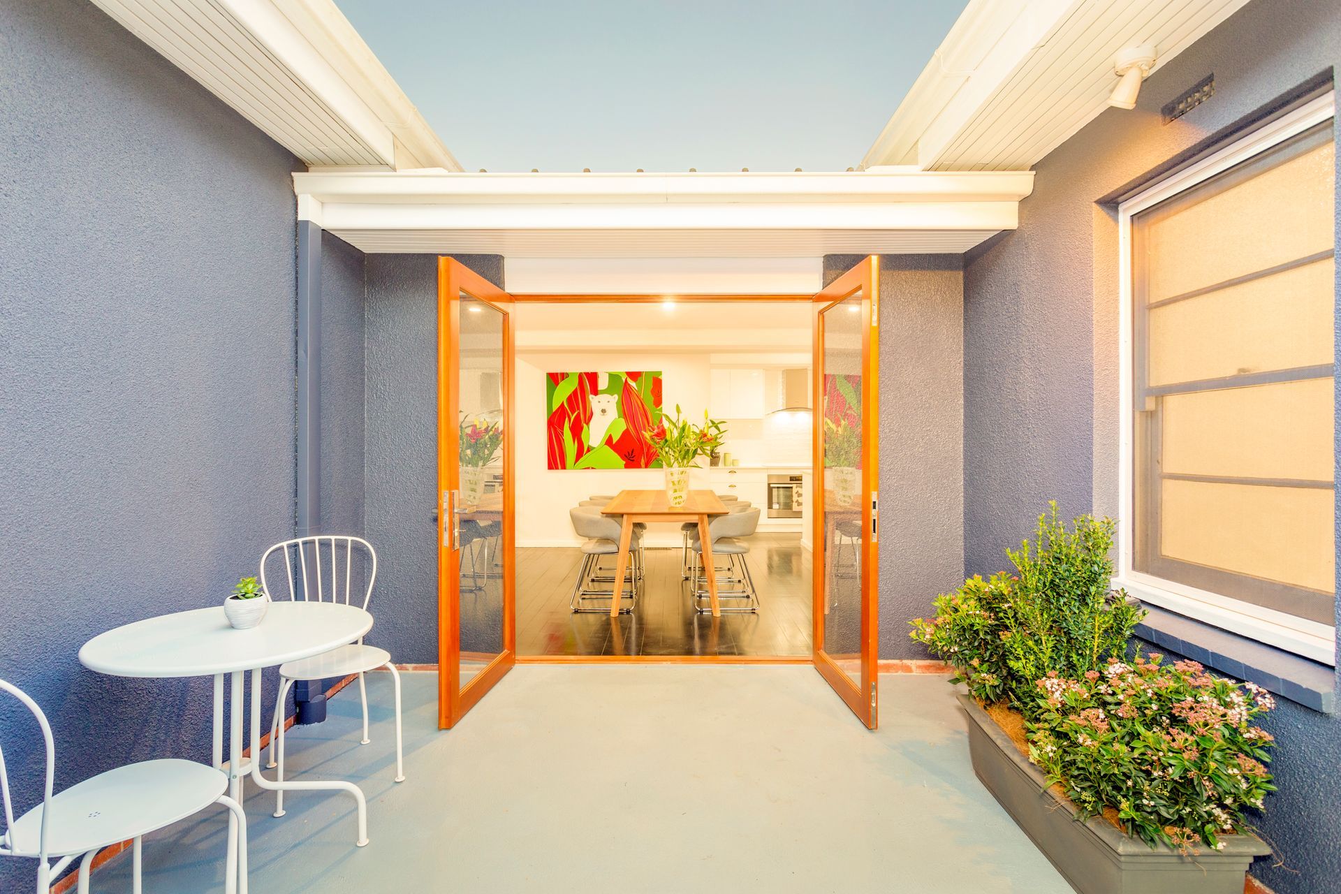 Patio with white table, chairs, and potted plants, opening to a dining room with a table and artwork.