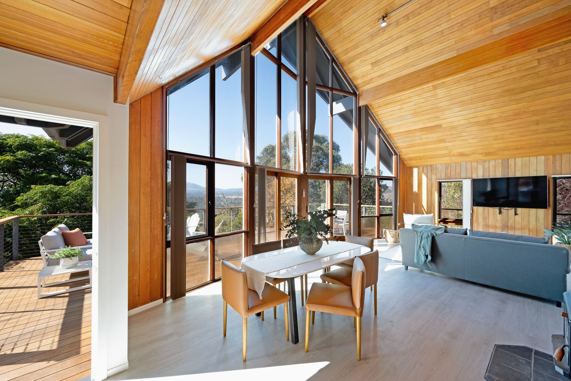 Living room with tall windows, wooden ceiling, dining table, and blue sofa, overlooking a scenic view.