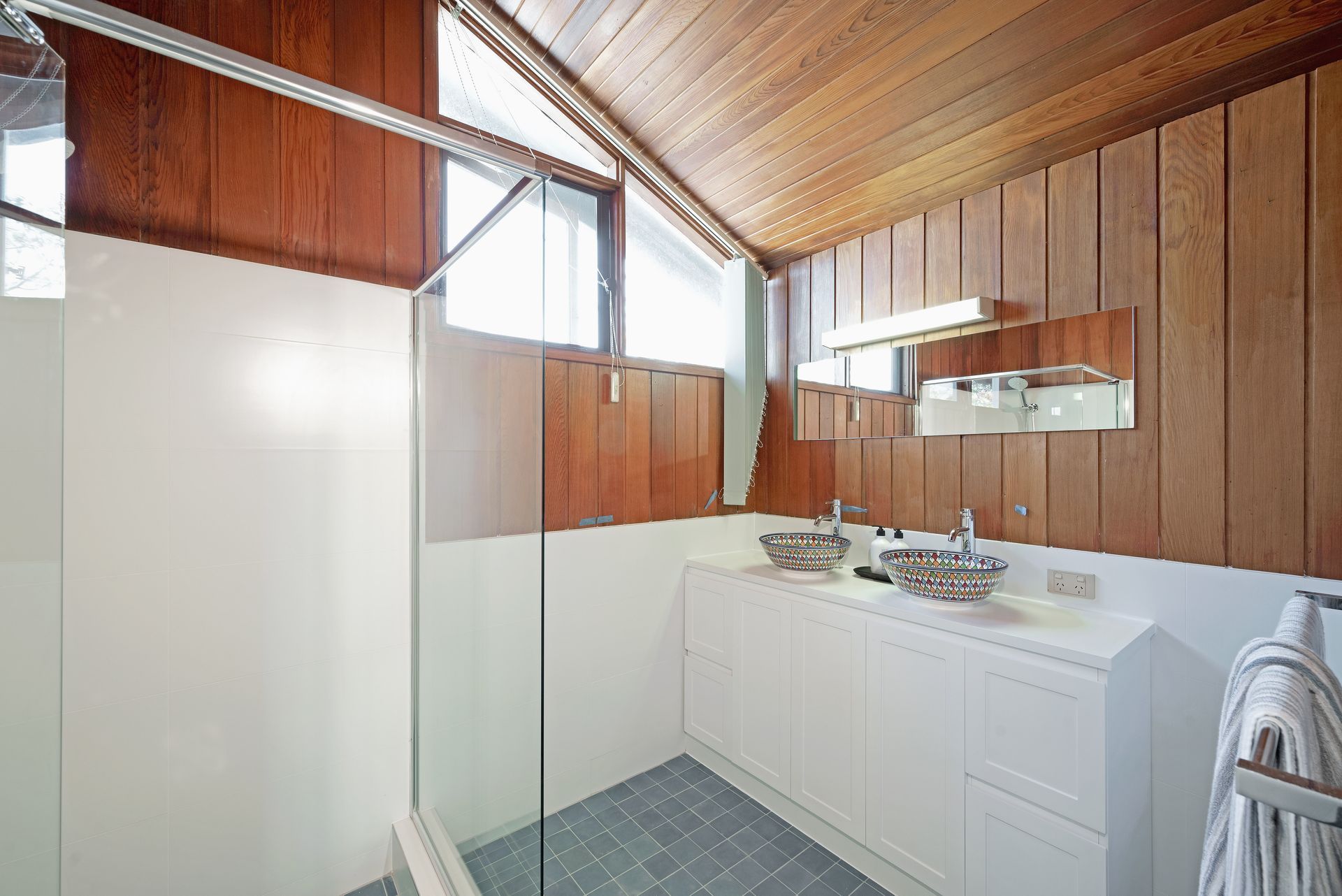 Bathroom with wood paneling and white cabinets. Glass shower door, two sinks with mirrors, blue tile floor.
