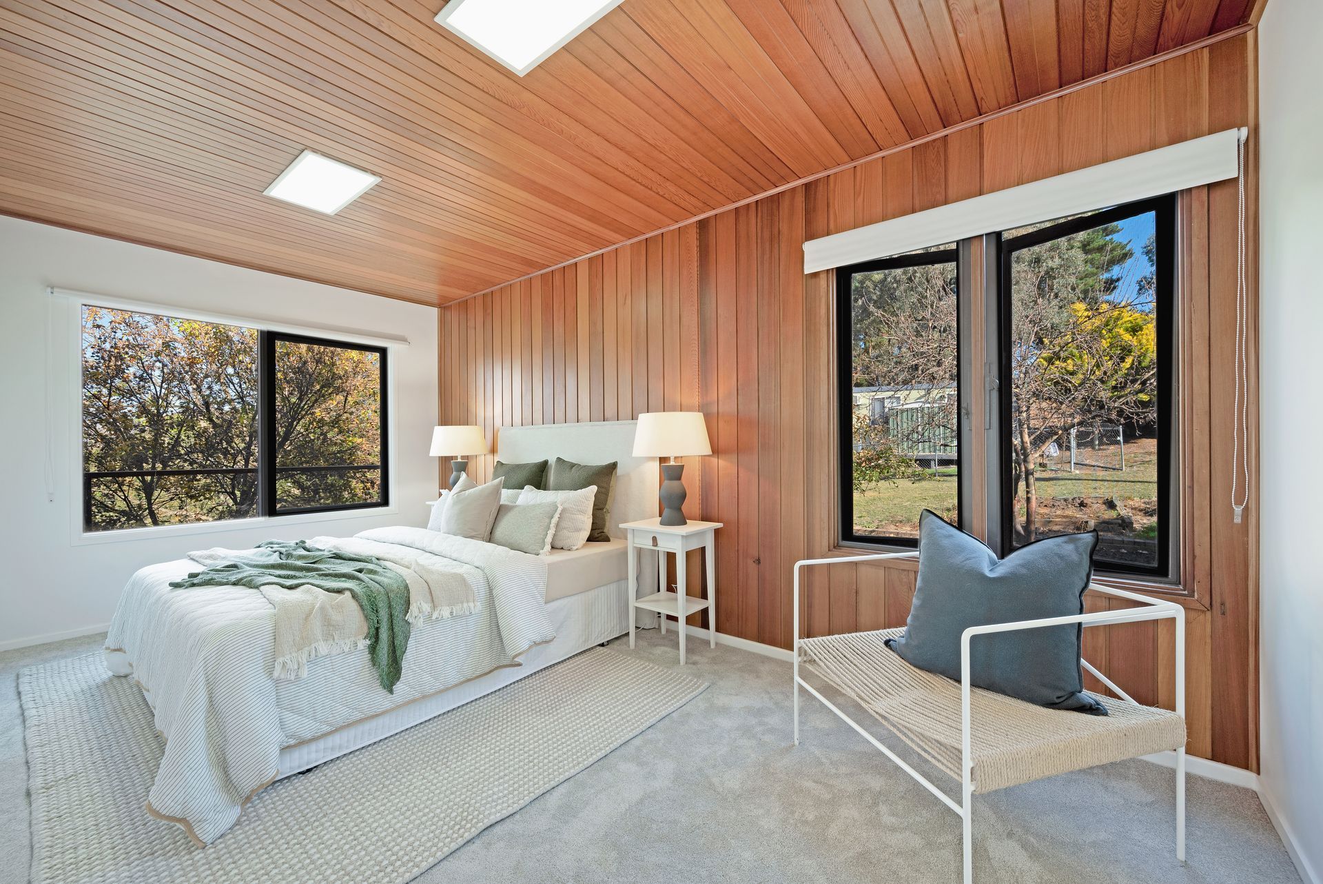 Bedroom with wood paneling, large windows, bed, bench, and two lamps.