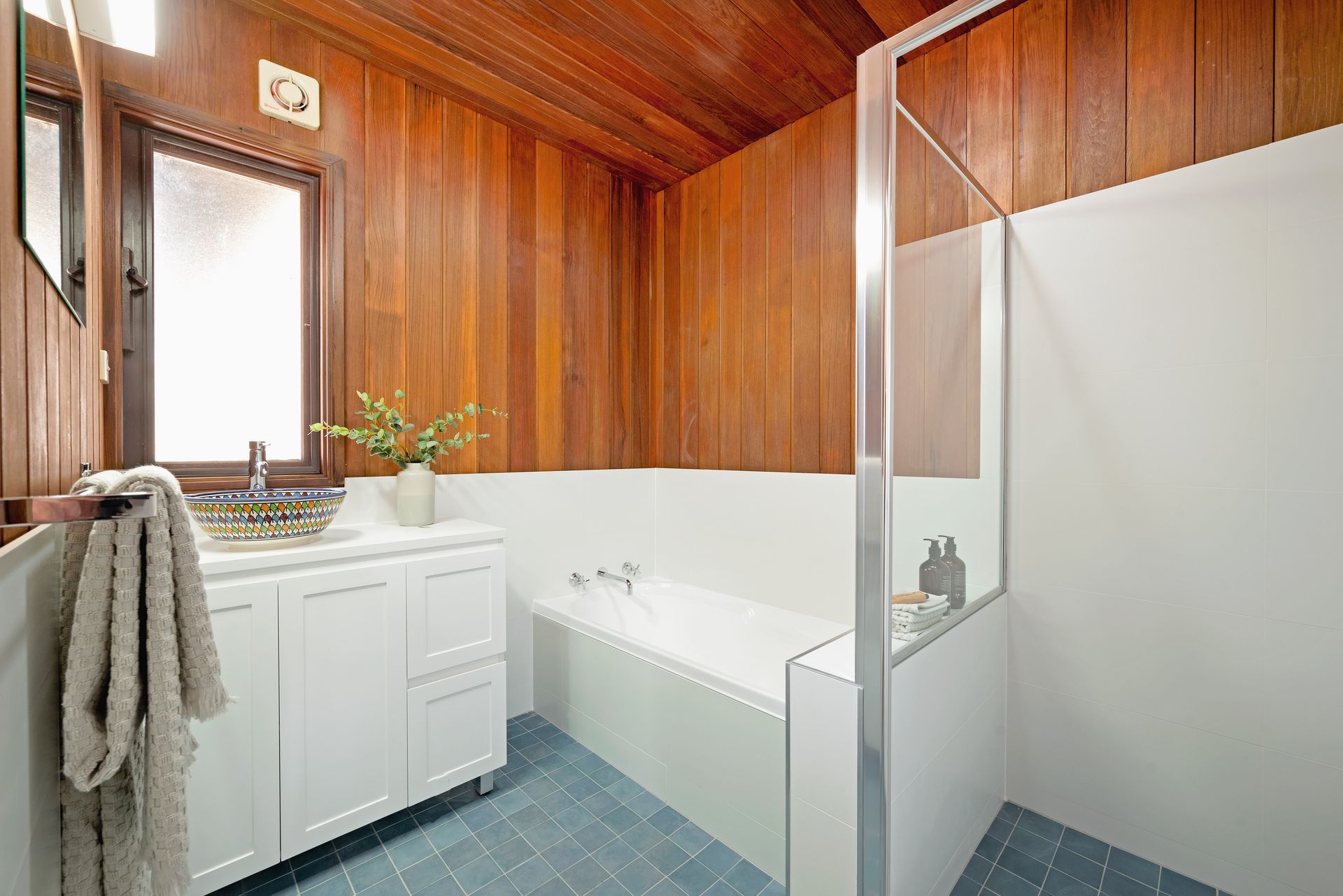 Bathroom with wood paneling, white tub and vanity, blue tile floor, and a glass shower screen.