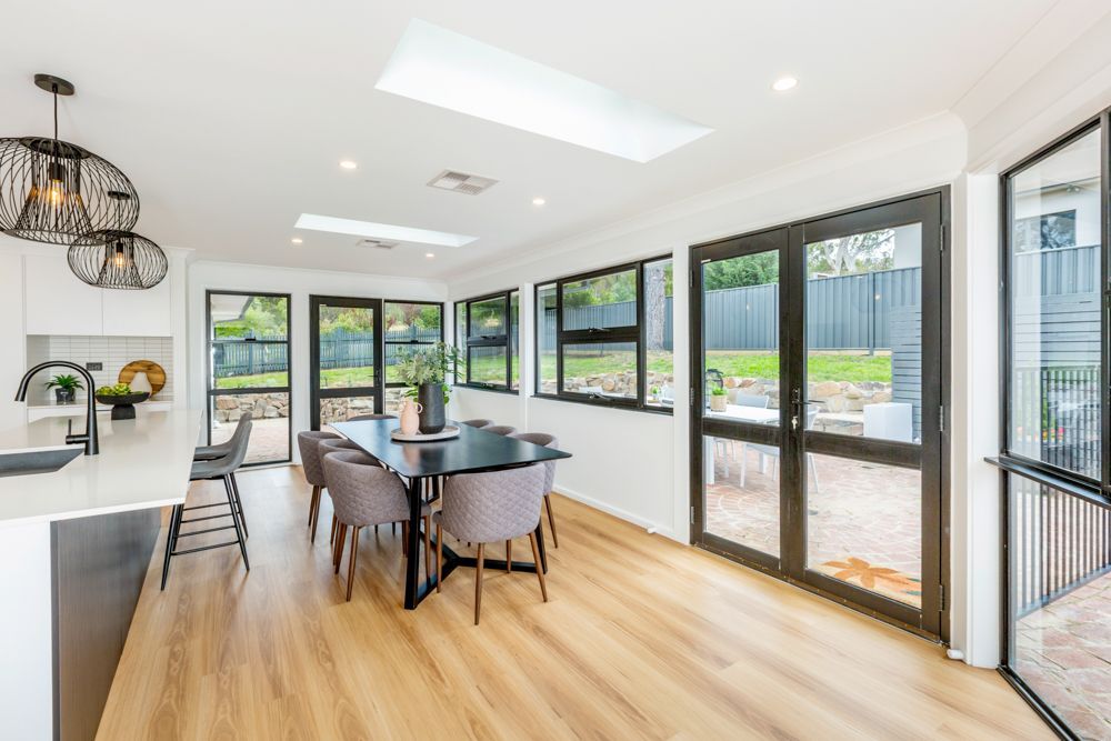 Dining room with black-framed windows, dark table, grey chairs, pendant lights, and light wood flooring.
