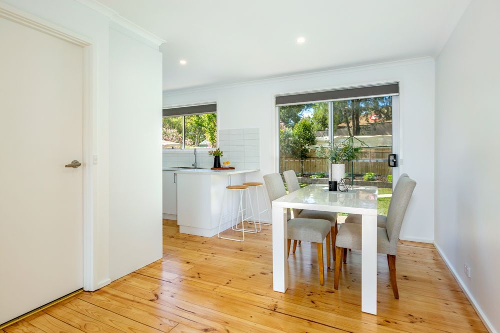Dining area with white table and chairs, adjacent to a kitchen and overlooking a garden through a window.
