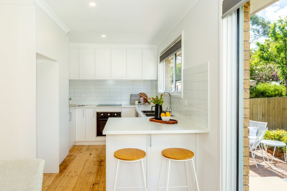 White kitchen with wooden floors, countertop with two stools, and a doorway to an outdoor patio with furniture.