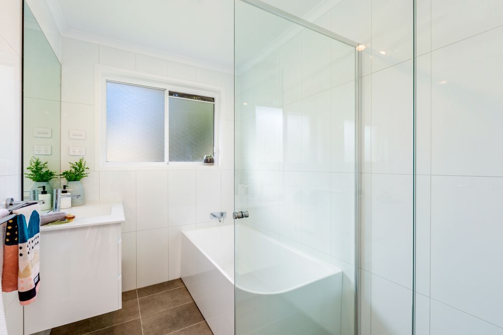 Bathroom with white tiles, glass shower, bathtub, and vanity with a window in the background.