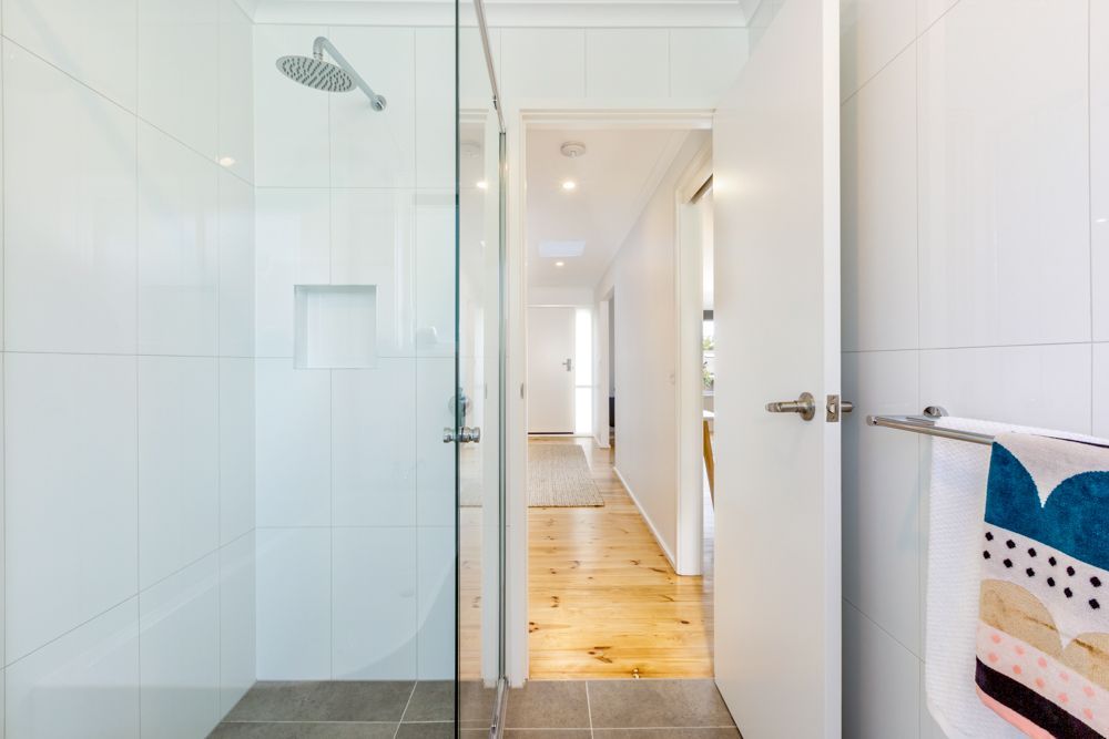 Bathroom with glass shower, white walls, and open door leading to hallway with wood floor.