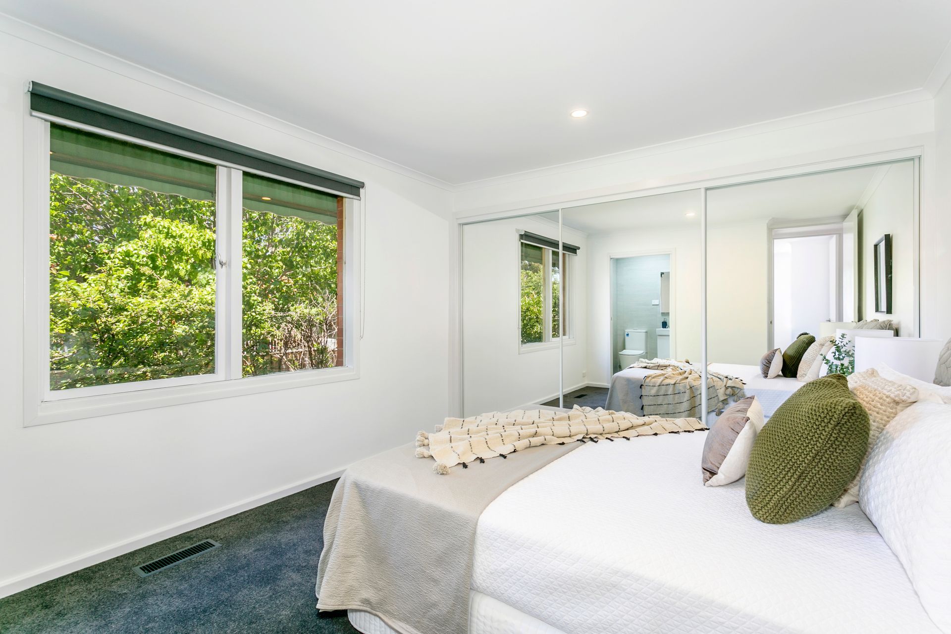 Bedroom with mirrored closet, window with green trees outside, and white bed with pillows.