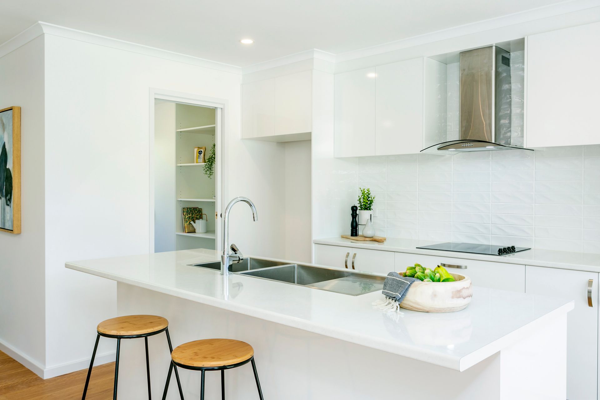 Modern white kitchen with island, stools, stainless steel range hood, and pantry in background.