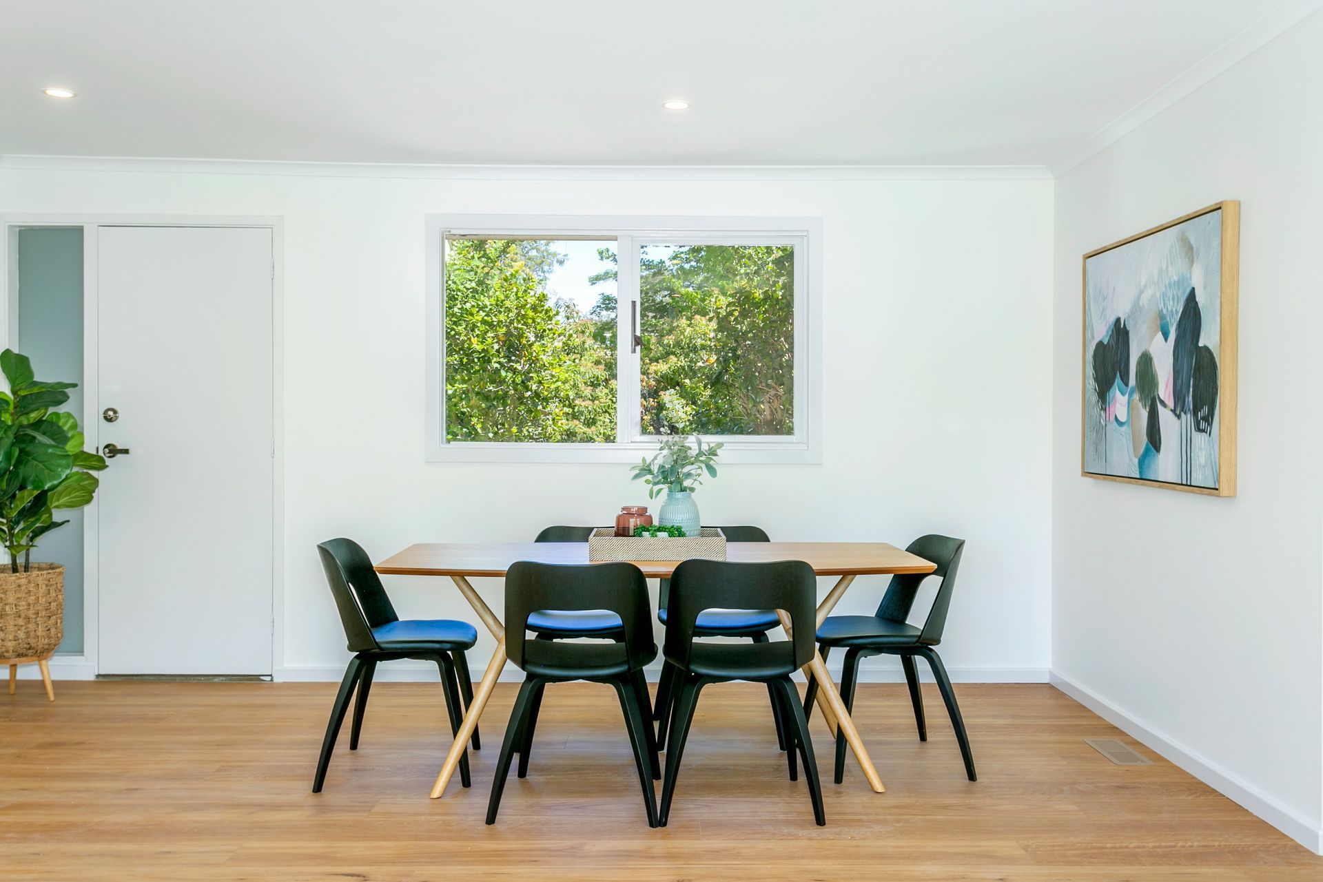 Dining room with wood table and black chairs, window overlooking greenery, art on the wall.