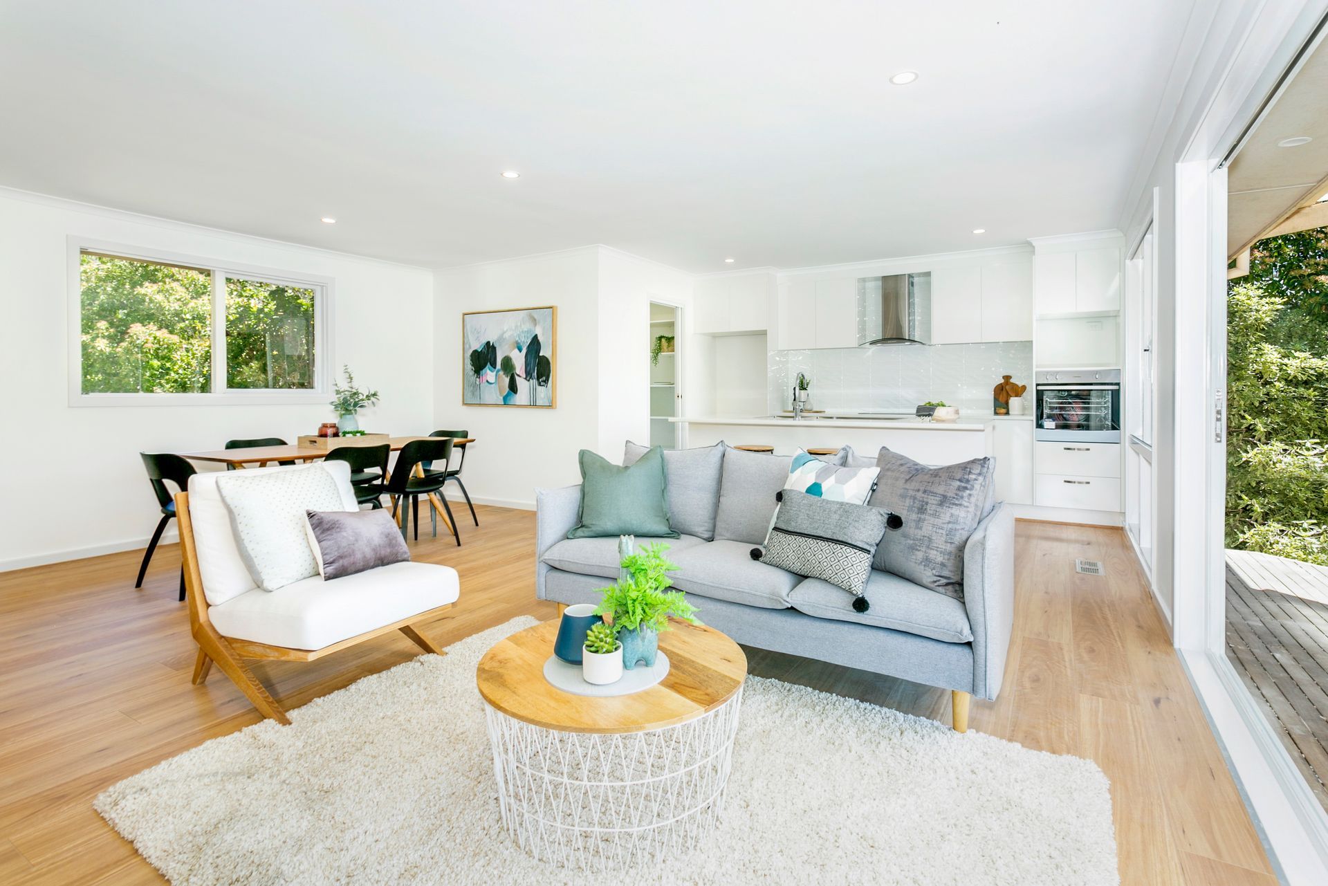 Living room with gray sofa, white armchair, wooden table, and rug. Open to kitchen and deck.