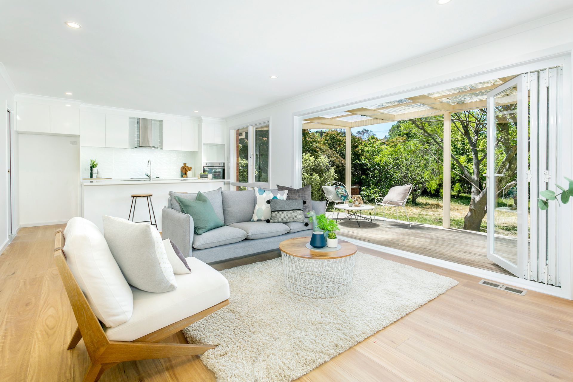 Bright living room with open doors to a backyard; light wood floors, gray sofa, white kitchen.