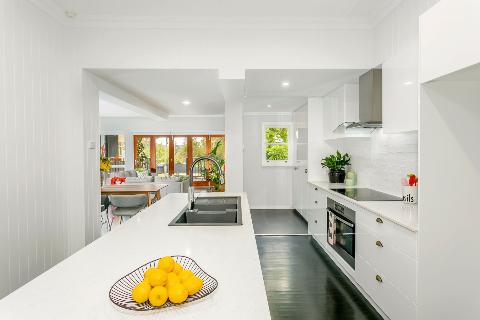 Modern white kitchen with island, black floors, and sunlight from a window.