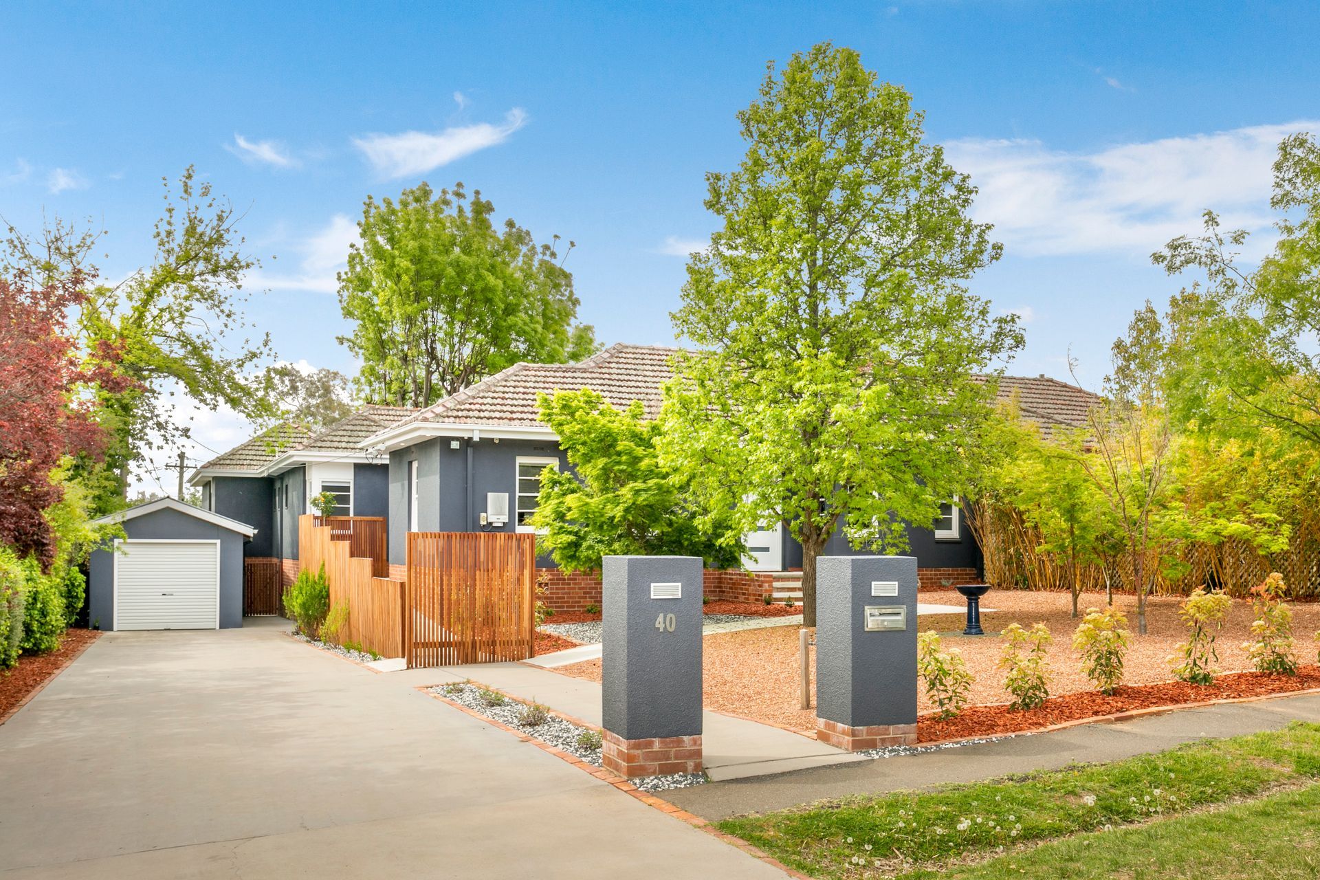 Gray house with attached garage, wooden fence, brick pillars, and trees. Sunny day.