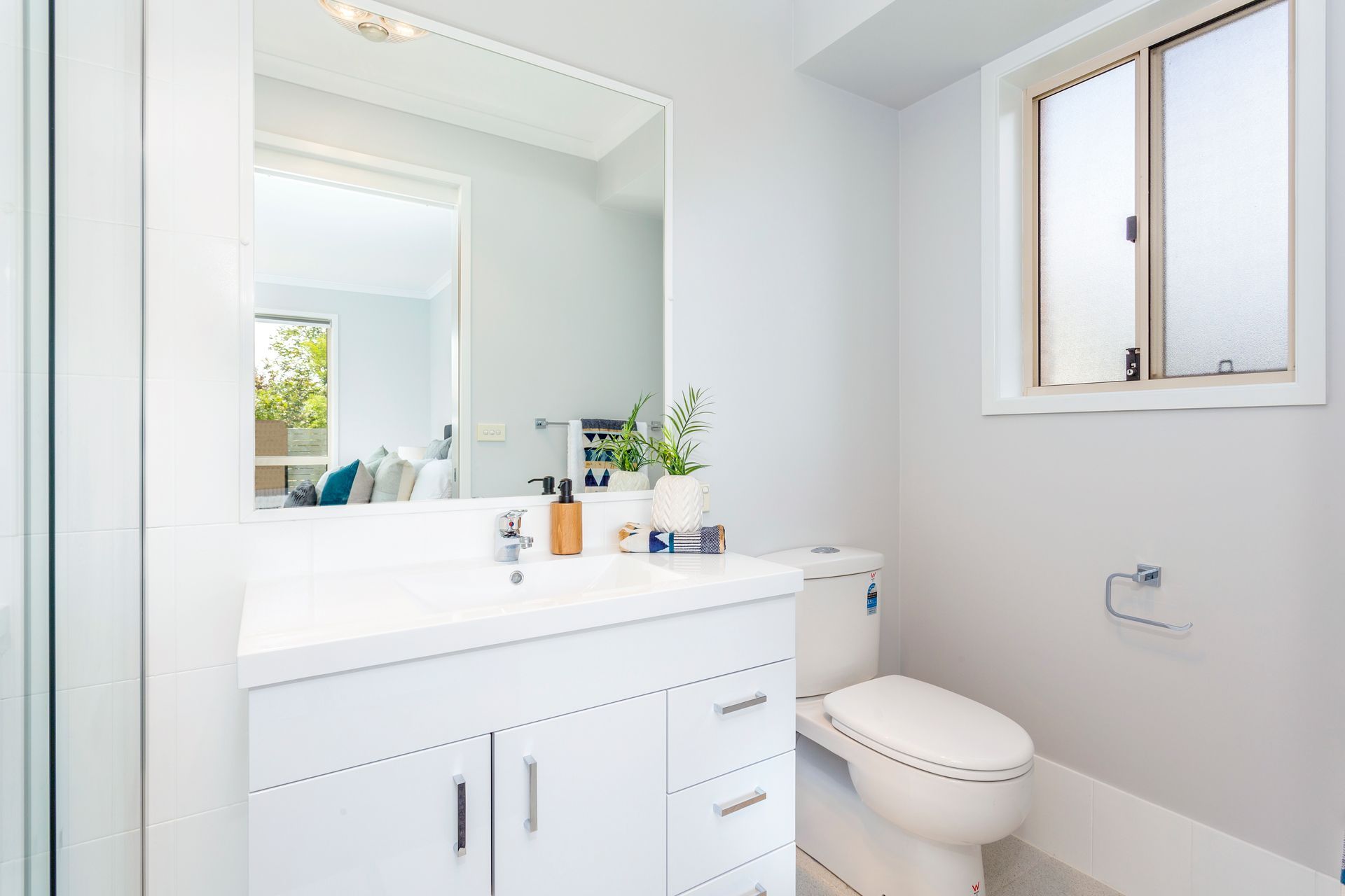 White bathroom with vanity, toilet, and window; mirror reflects partial view of a living room.