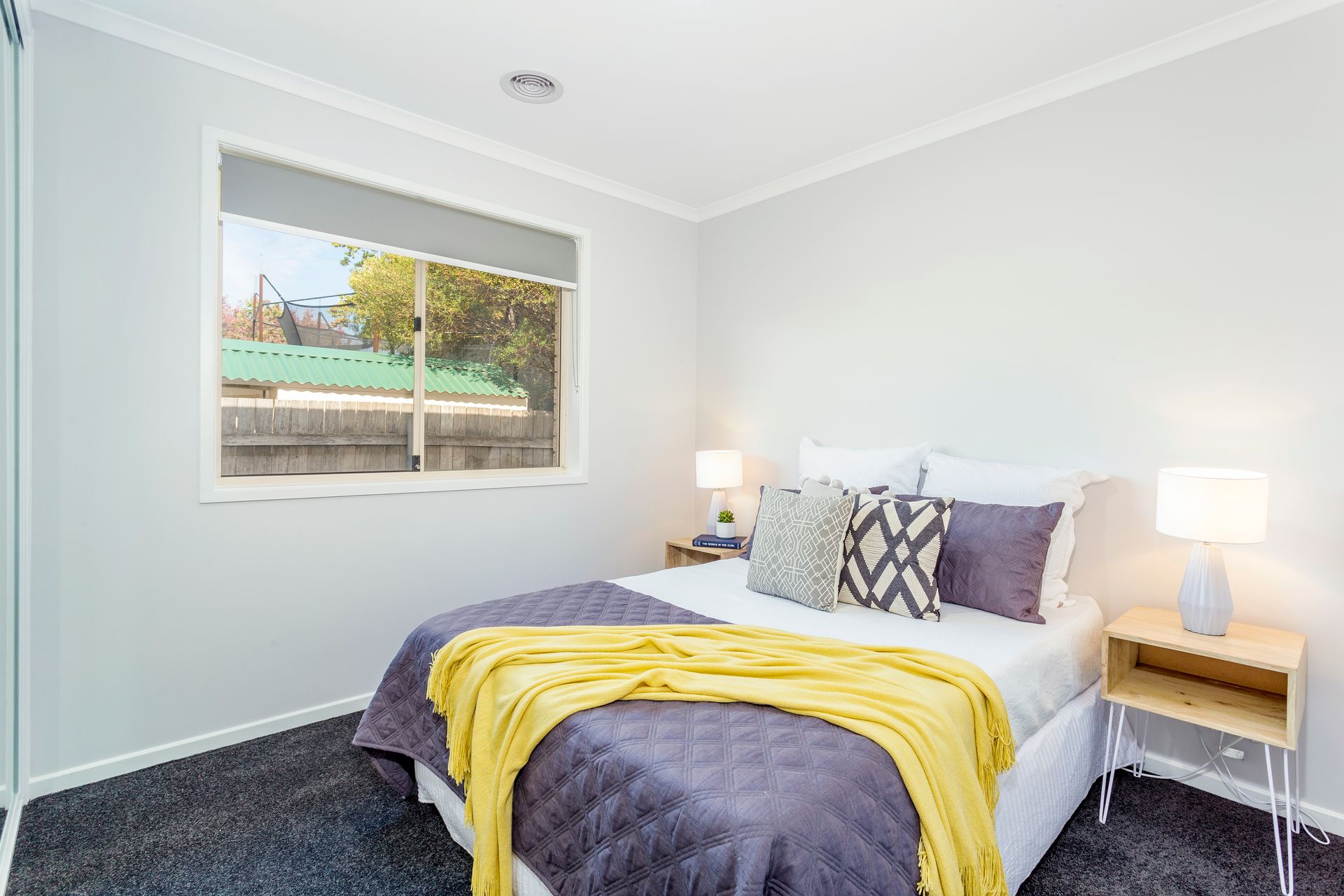 Bedroom with gray walls, bed with purple quilt and yellow blanket, window, and two bedside tables.