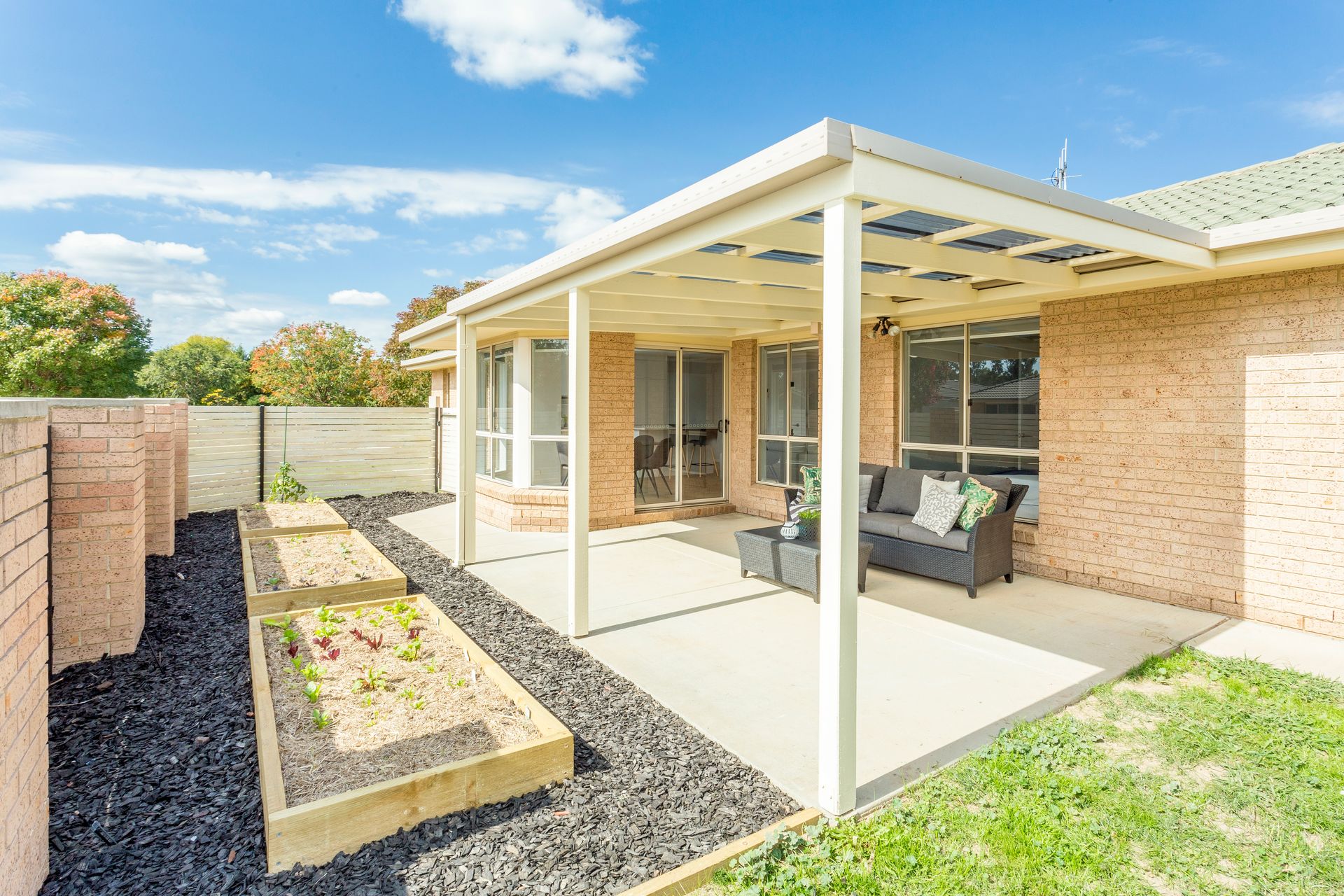 Covered patio with seating, raised garden beds, and brick wall. Sunny day, blue sky.