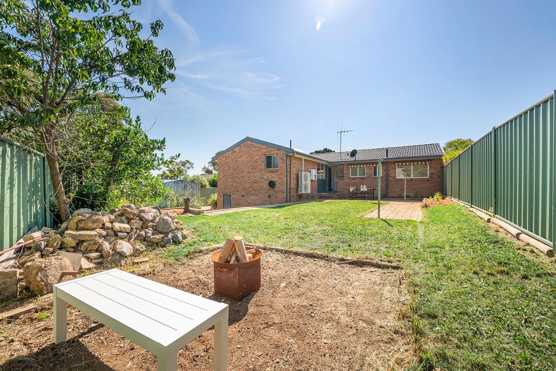 Backyard with lawn, brick house, white bench, fire pit, and green fences under a sunny sky.
