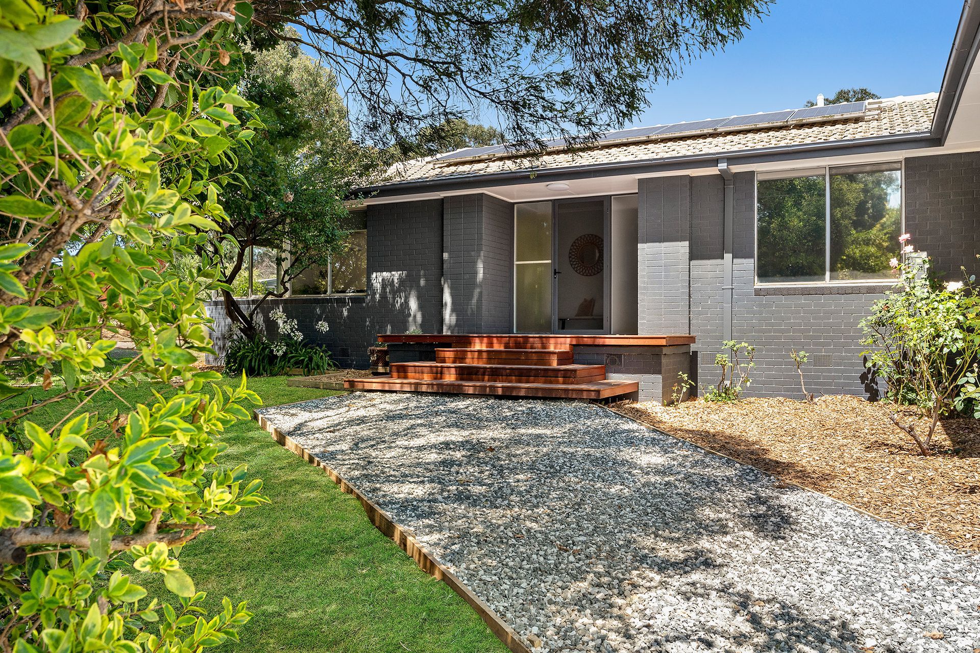 House exterior with gray facade, wooden steps, gravel path, and greenery.