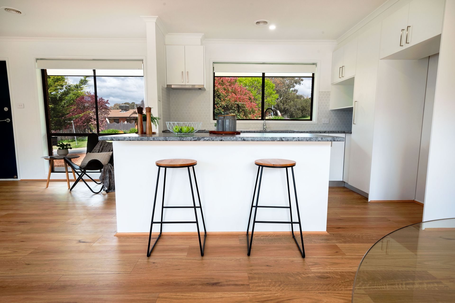 Modern kitchen with island, two stools, wooden floors, and large windows with a view.
