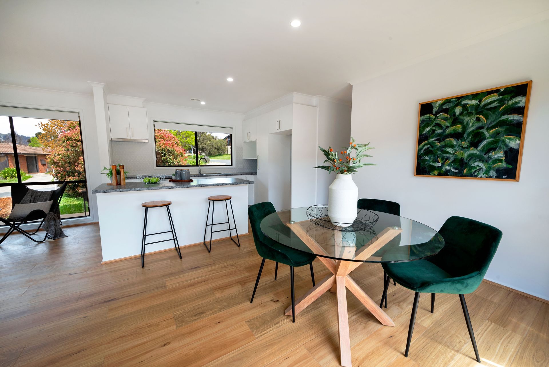 Dining area with glass table, green chairs, and artwork, connected to a kitchen with a breakfast bar.