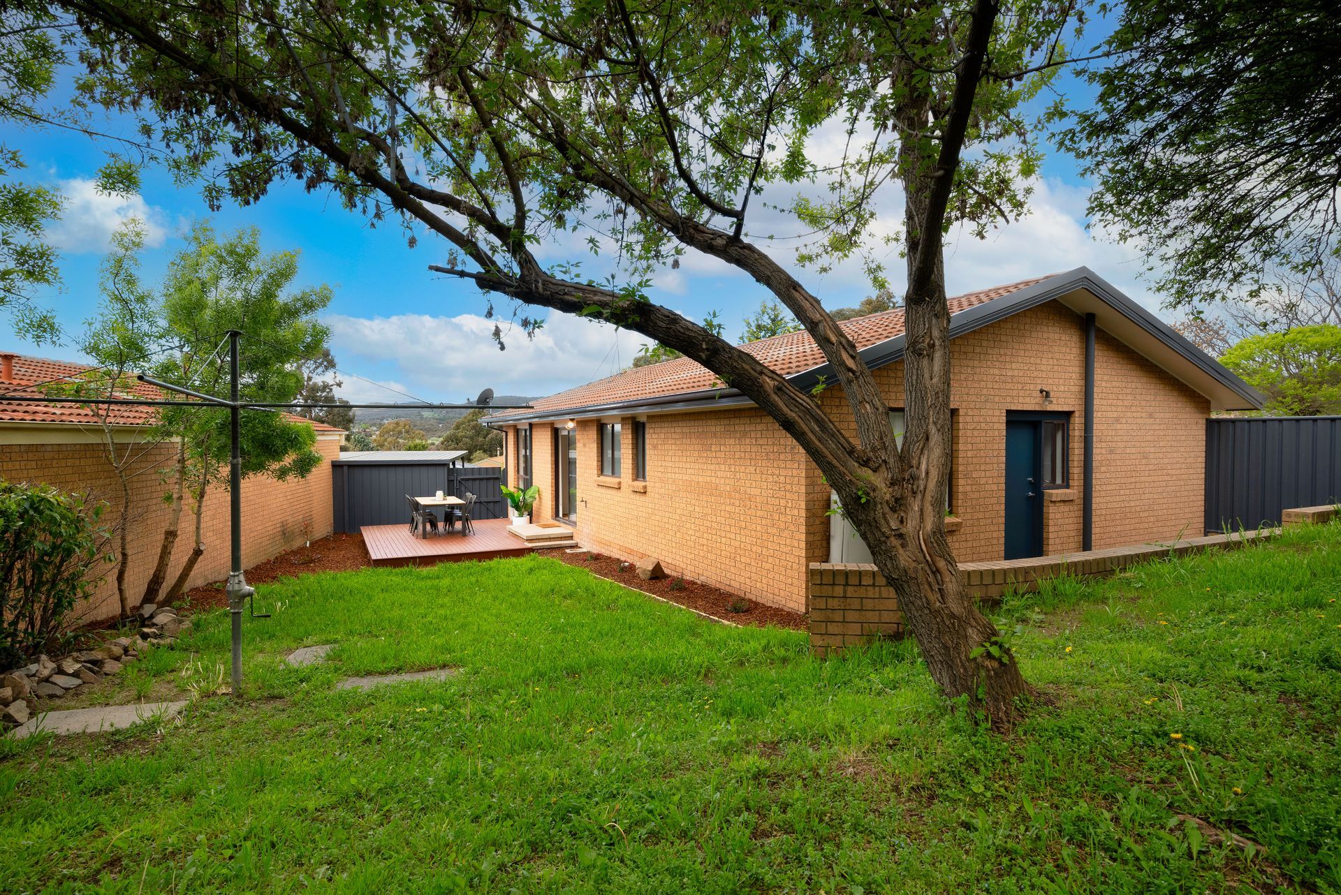 A brick house with a green lawn, tree branches, and a small patio with a table.
