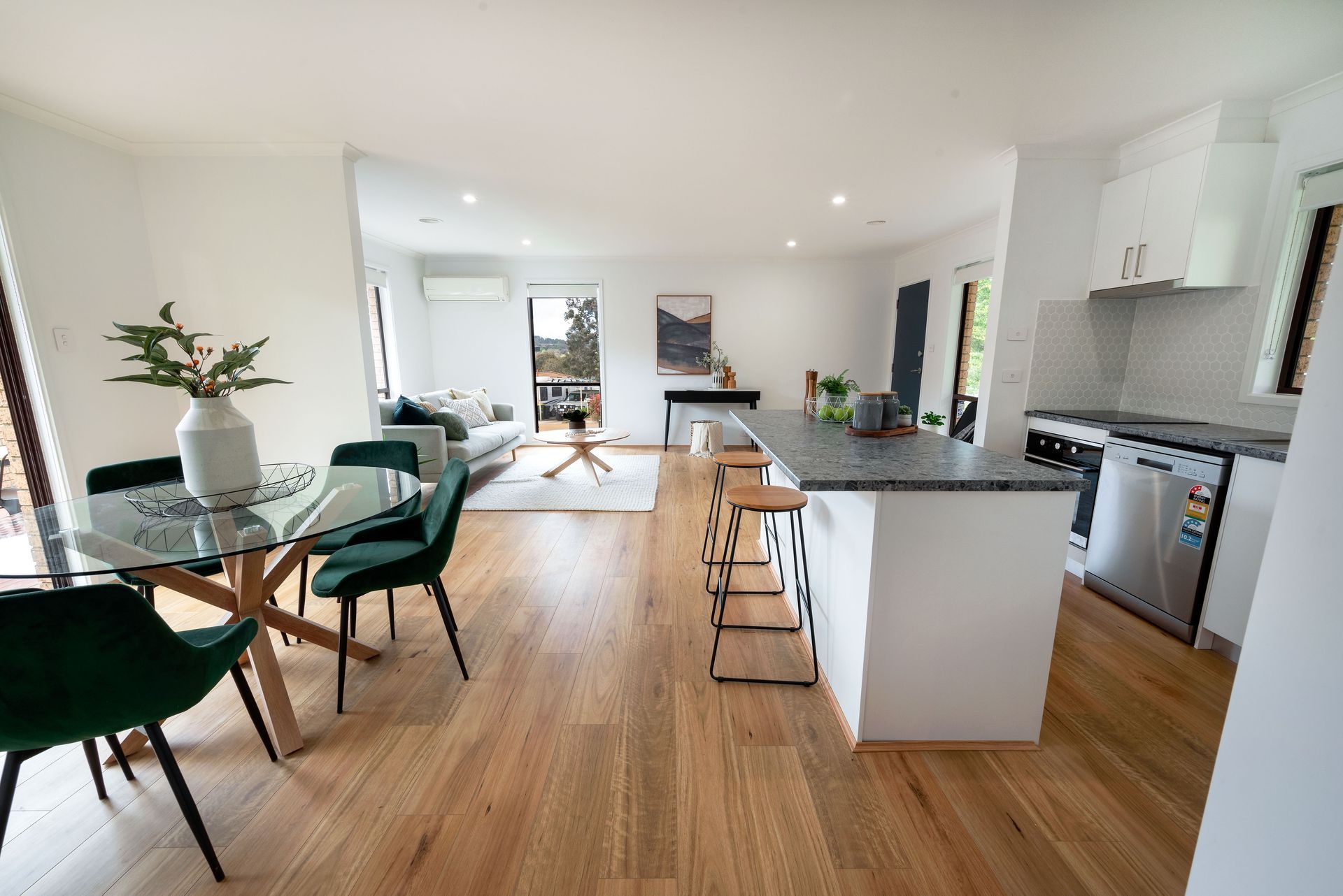 Open-plan living space with kitchen island, dining table, and living area with couch, wooden floors, and white walls.