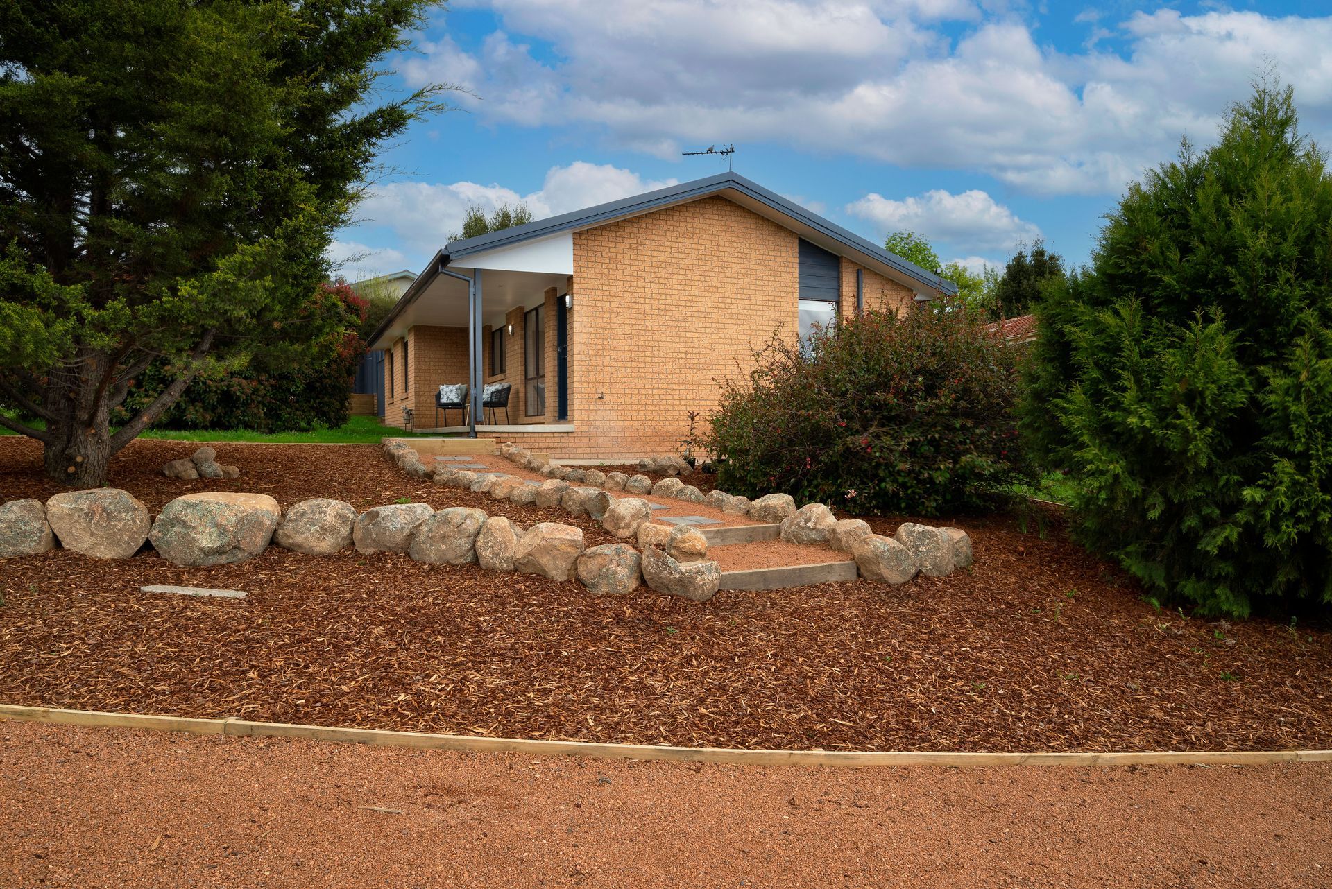 Small brick house with a front porch, surrounded by trees and a mulch pathway leading up to the entrance.