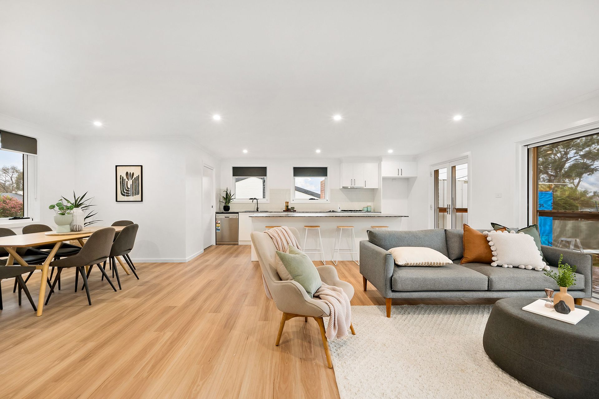 Open-plan living area with dining table, kitchen, and sofa. Light wood floors, white walls, and neutral furniture.