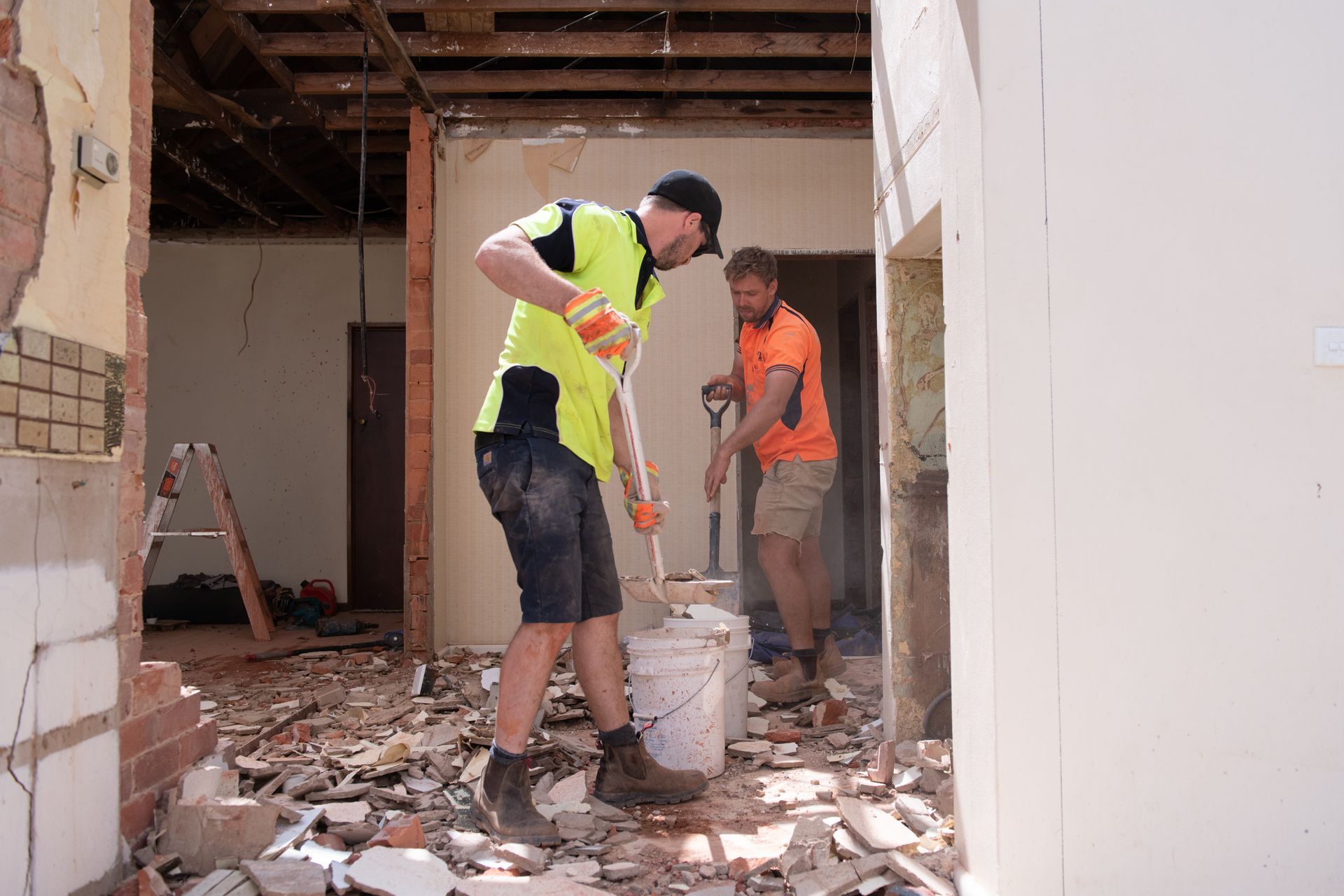 Two construction workers clearing debris during a demolition. One wearing a yellow vest scoops into a bucket.