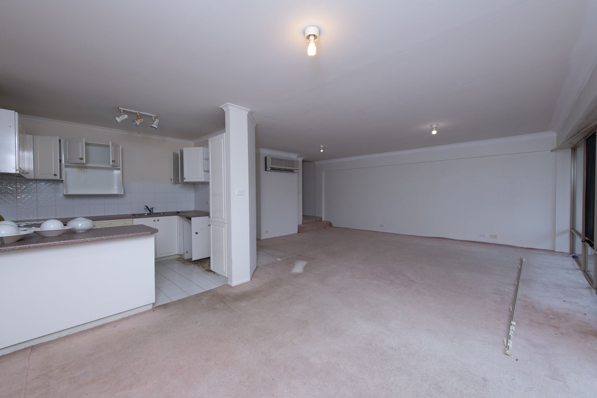 Empty, open-plan living space with kitchen area, white walls, and worn carpet.