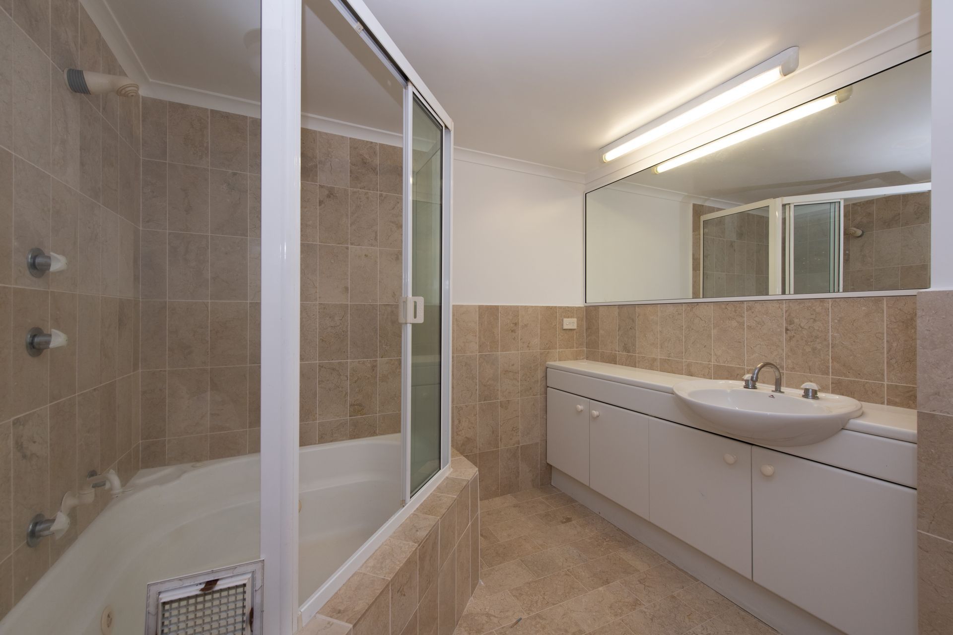 Bathroom with a tub/shower combo. Beige tile, white vanity, large mirror, and glass shower door.