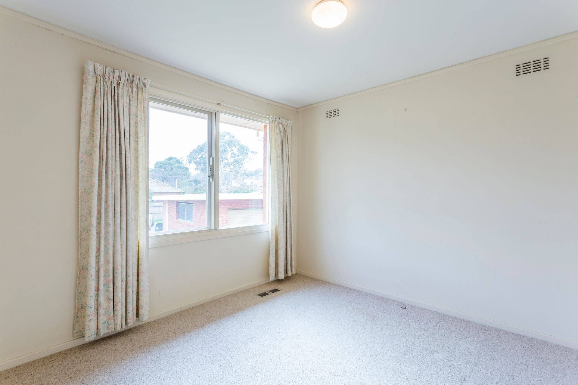 Empty room with a window, beige walls, and light carpet. White curtains frame the window.