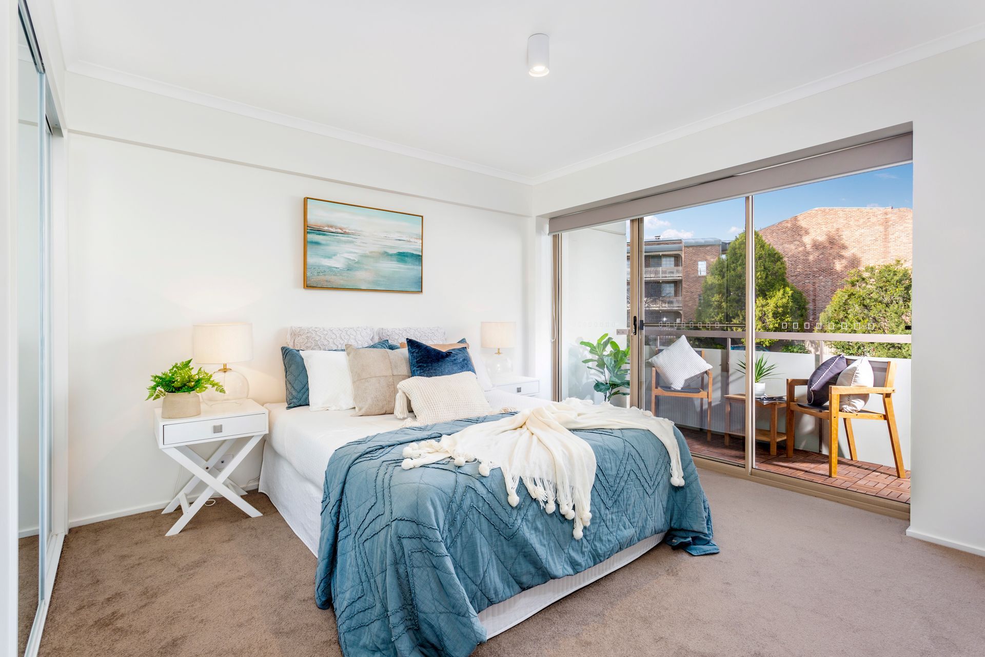 Bedroom with bed, balcony, and white walls, featuring blue bedding and artwork.